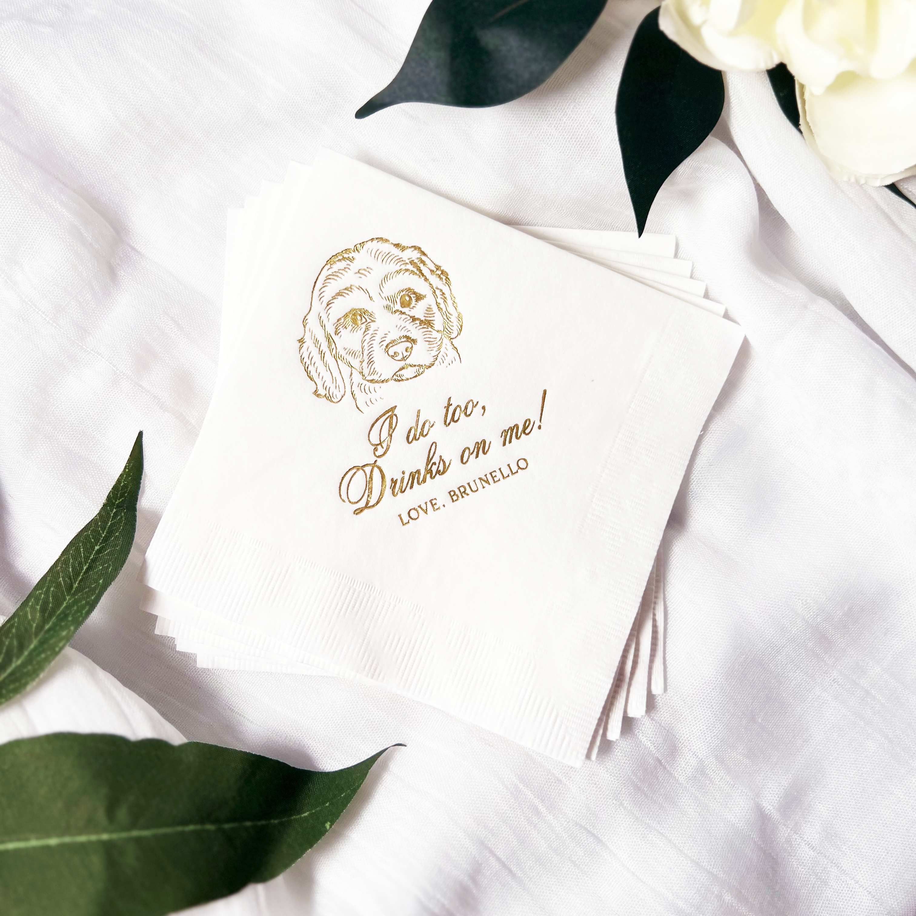 White paper napkins featuring a custom gold foil dog illustration and a personalized wedding-themed message, styled on a textured white fabric with greenery and white flowers.