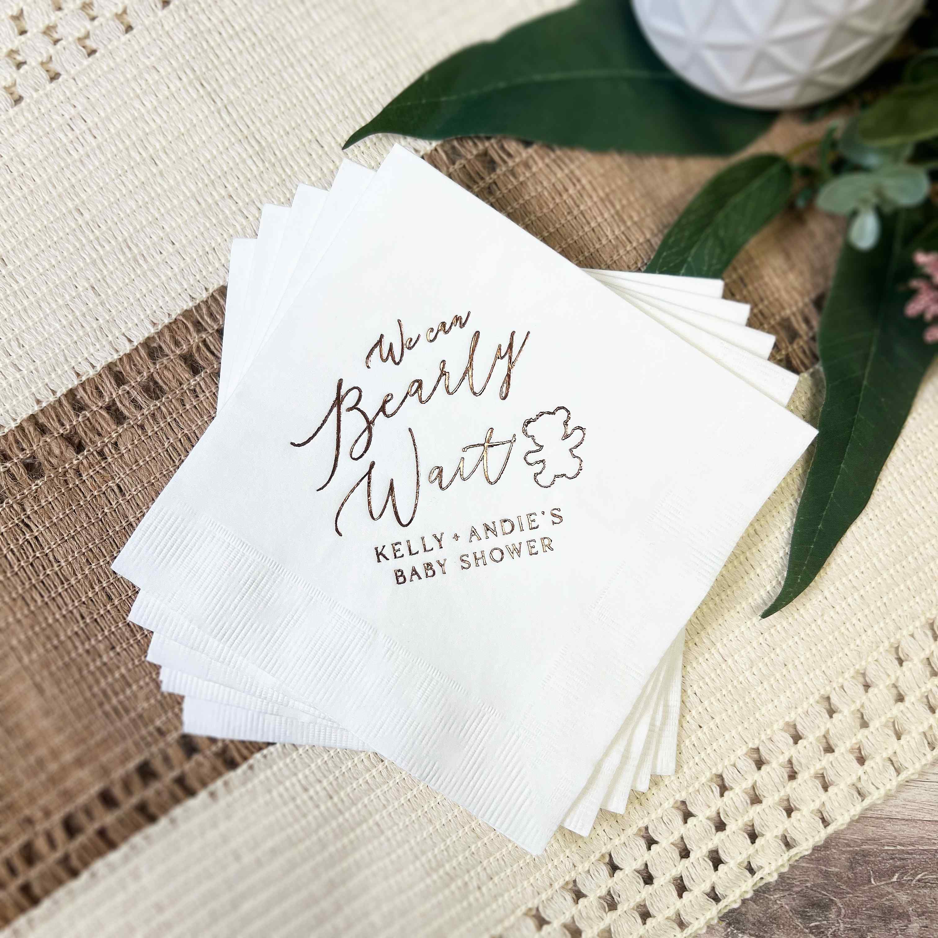Stack of white paper beverage napkins featuring rose gold foil lettering that reads We can Bearly Wait with a cute bear cub illustration, ideal for a baby shower celebration.