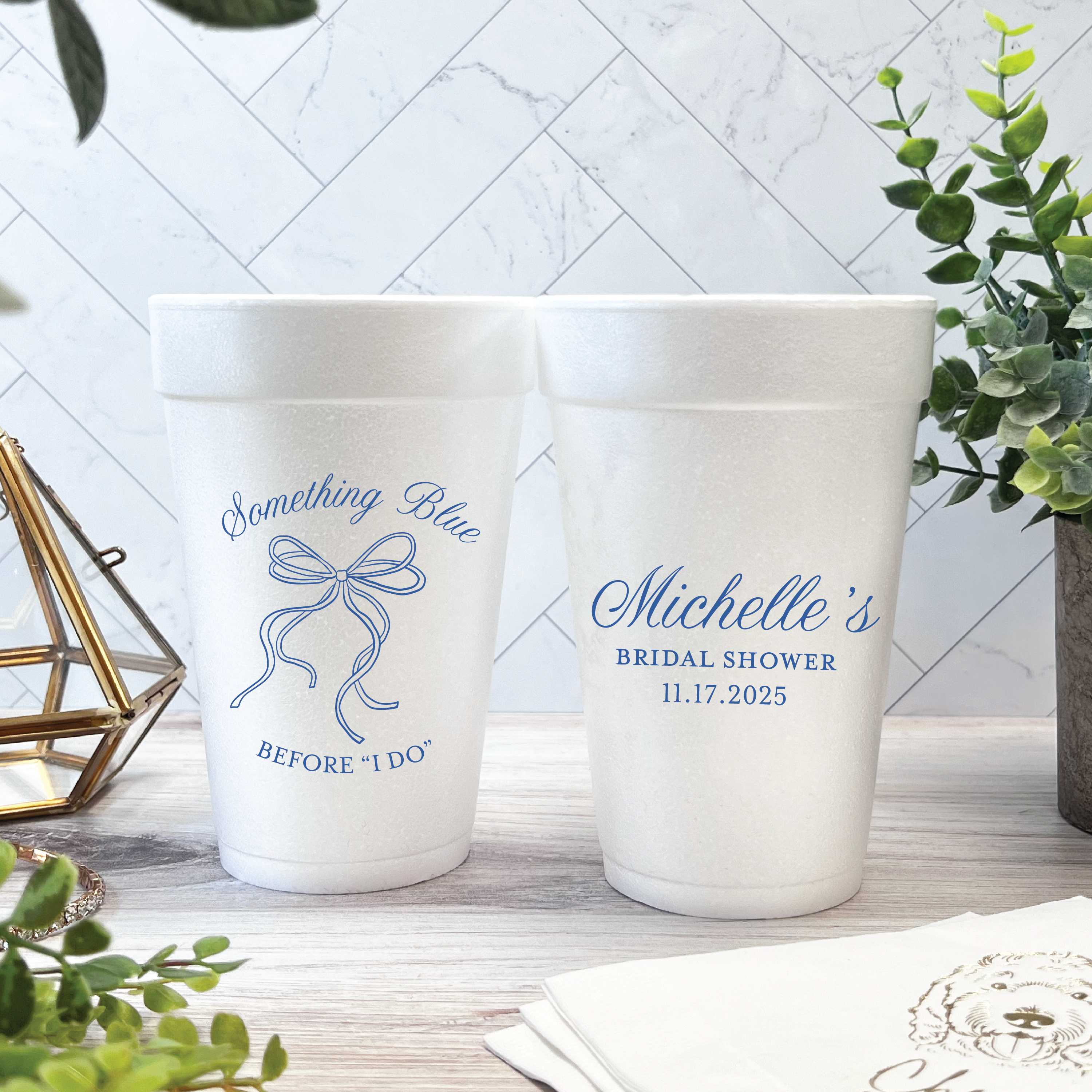 Custom white foam cups featuring a Something Blue bow design and personalized bridal shower text, arranged on a rustic wooden surface with decorative greenery.
