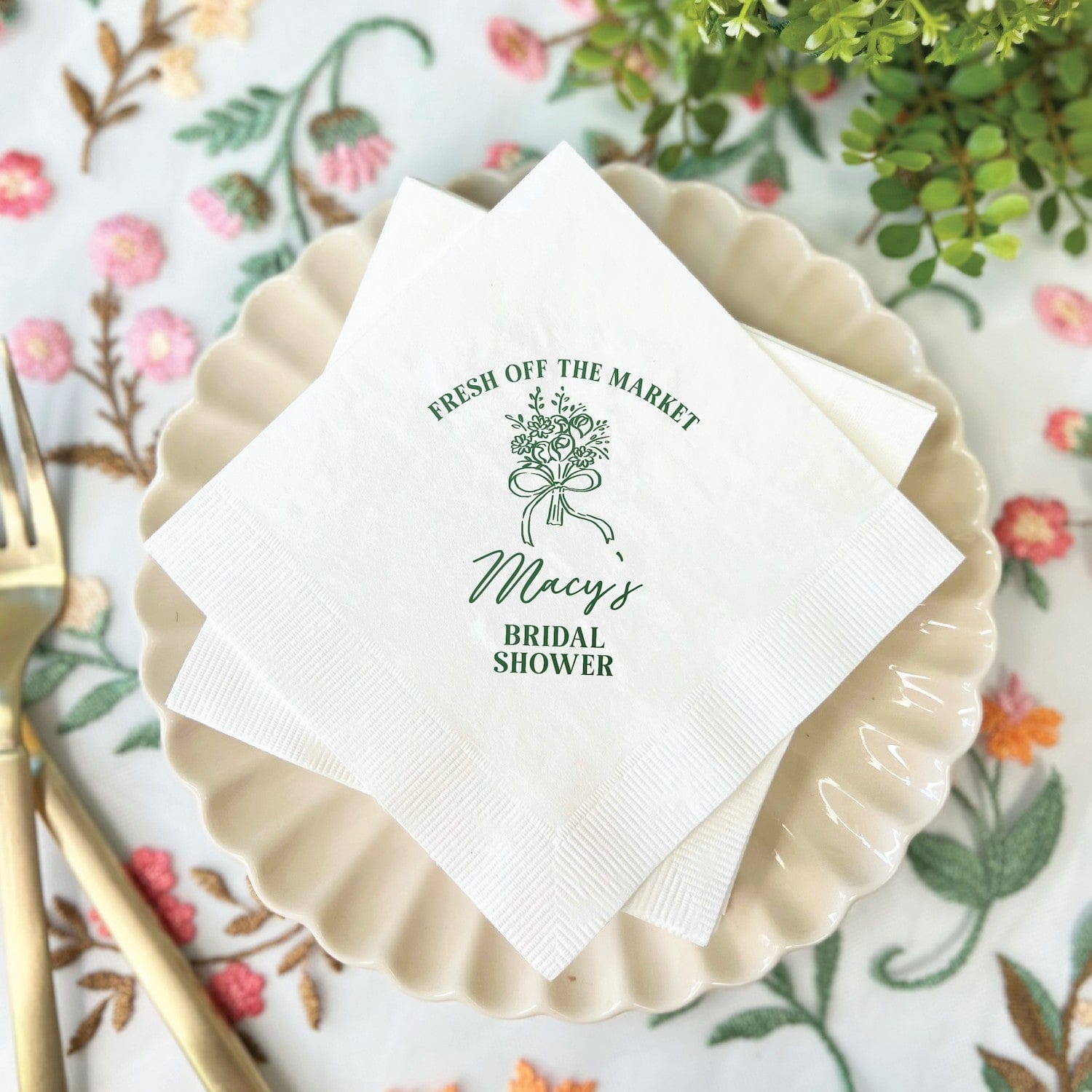 Personalized paper napkins for a bridal shower featuring a Fresh Off The Market design with a bouquet graphic printed in green ink, set on a scalloped plate with a floral tablecloth.