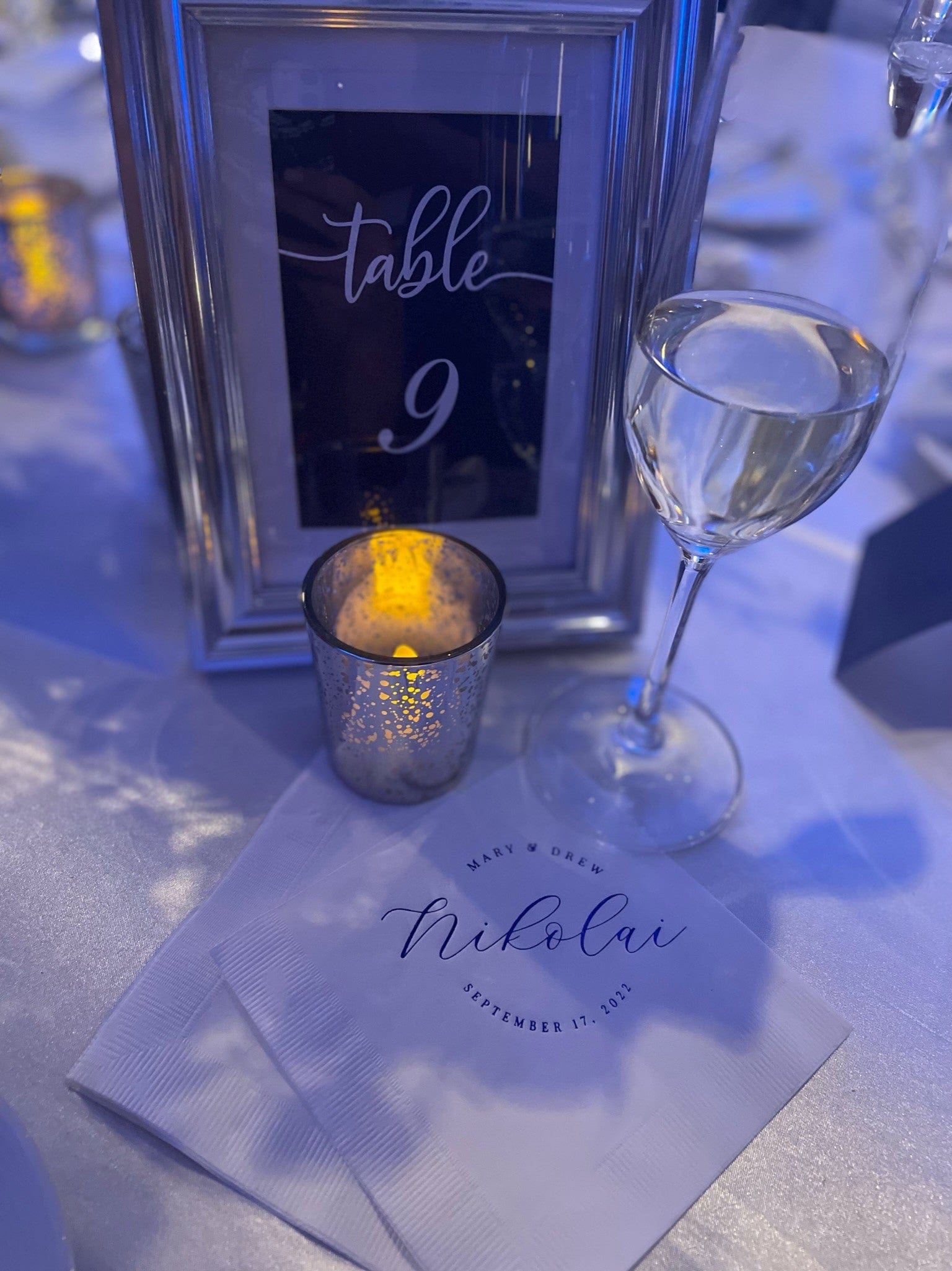 A personalized wedding napkin with custom script text elegantly placed on a white tablecloth as part of a formal dinner table setting, alongside a framed table number and glowing votive candle.