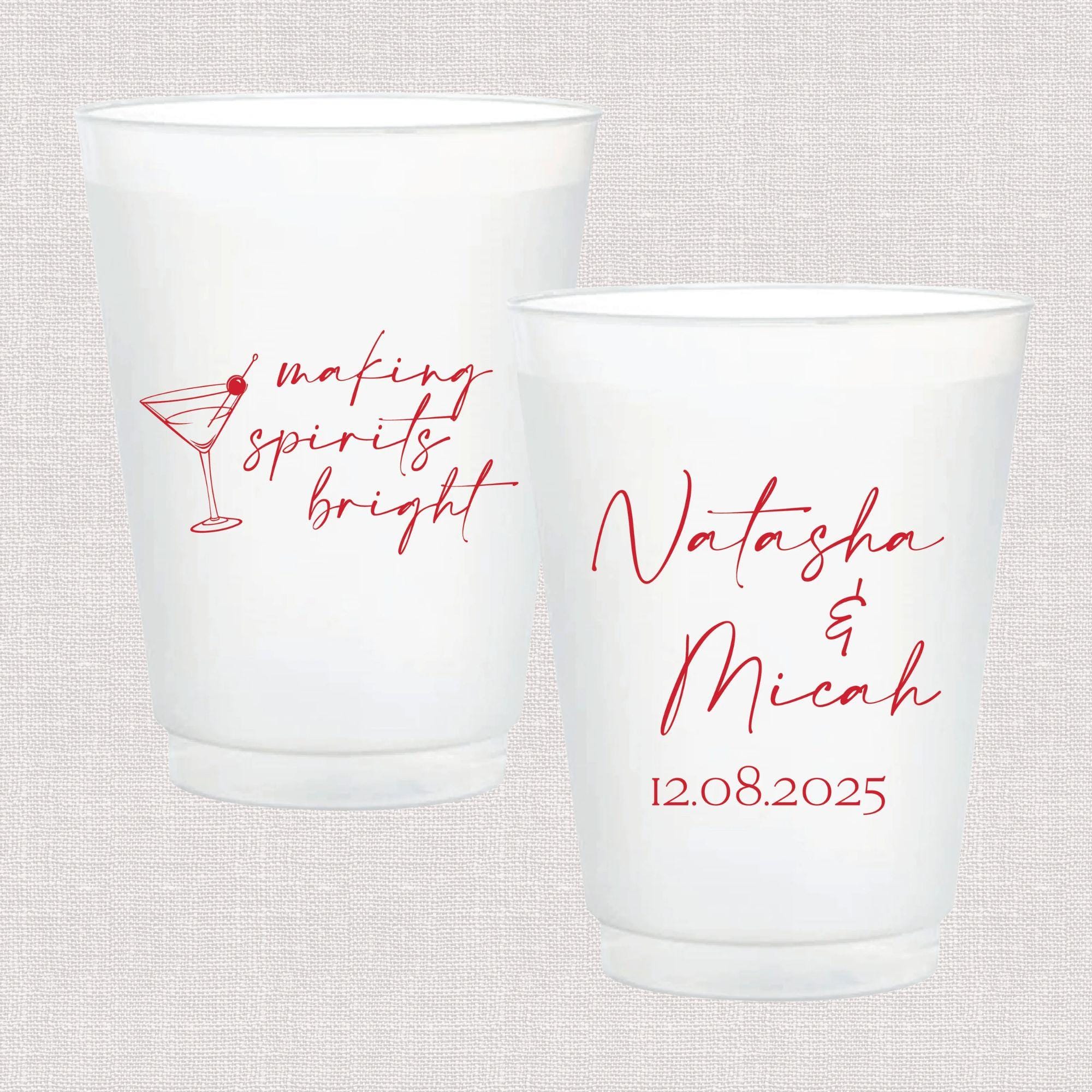 Two frosted plastic cups with red script text, one featuring a cocktail glass and "making spirits bright," the other personalized with names "Natasha & Micah" and date "12.08.2025"