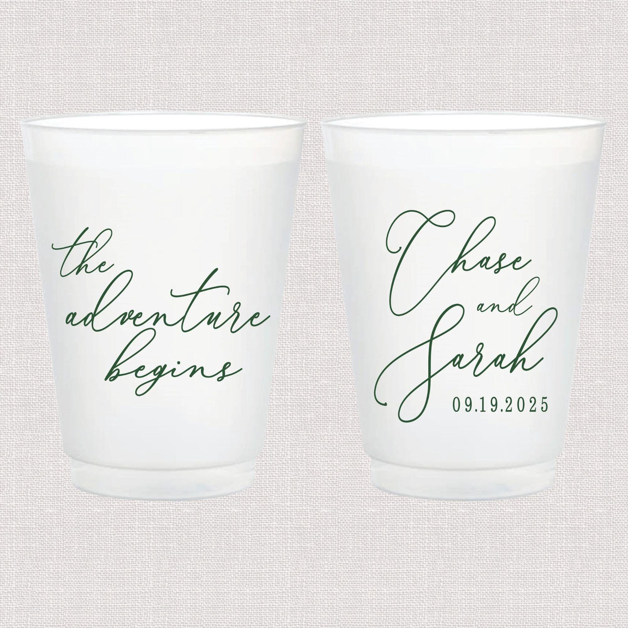 Pair of white frosted cups with cursive text, one reading "the adventure begins" and the other personalized with names and wedding date