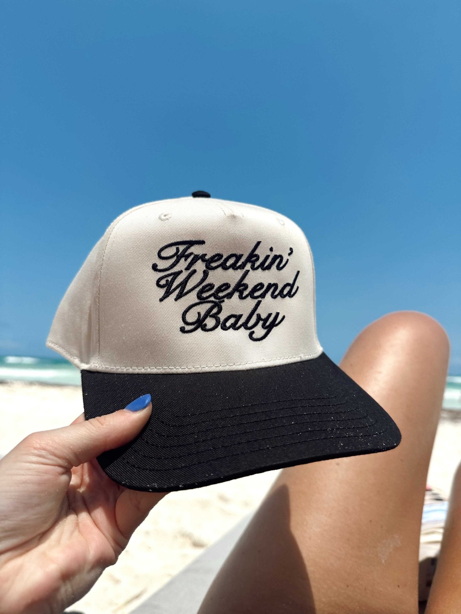 Cream and black vintage trucker hat with "Freakin' Weekend Baby" embroidered text, held up at the beach.