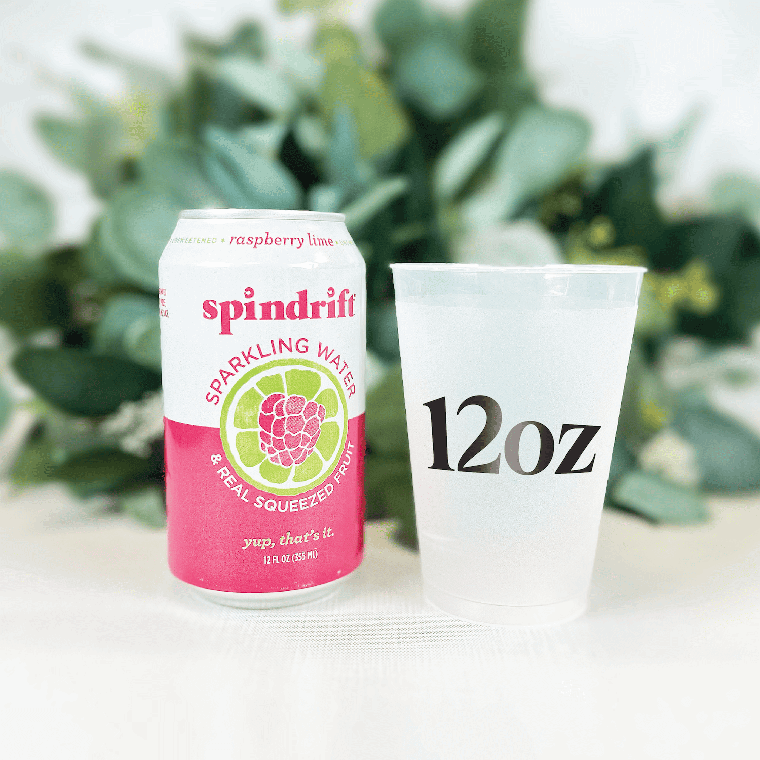 White frosted 12oz cup next to pink Spindrift sparkling water can with lime slice design and greenery backdrop.