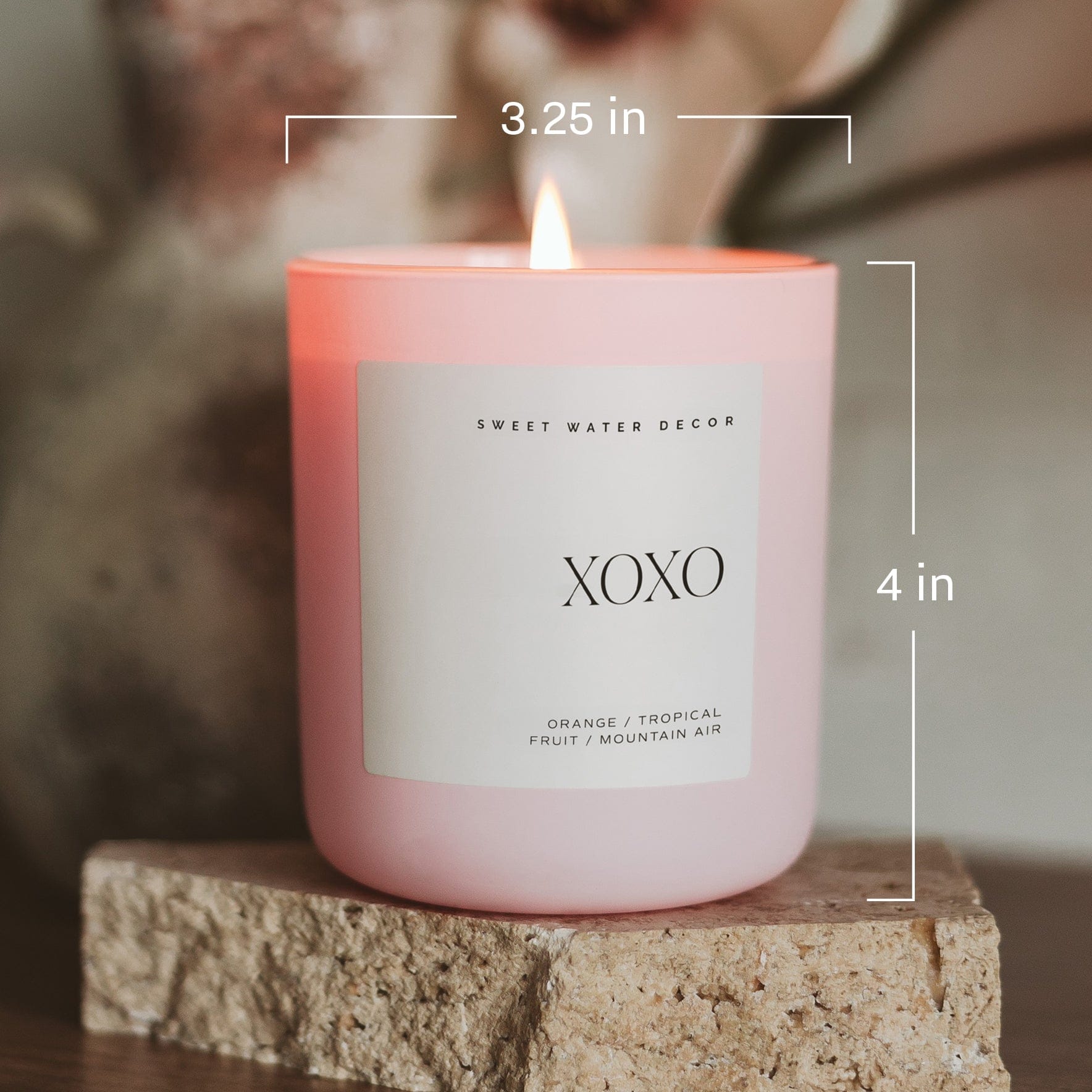Pink matte jar candle labeled XOXO with white label, burning flame on top, sitting on textured stone block