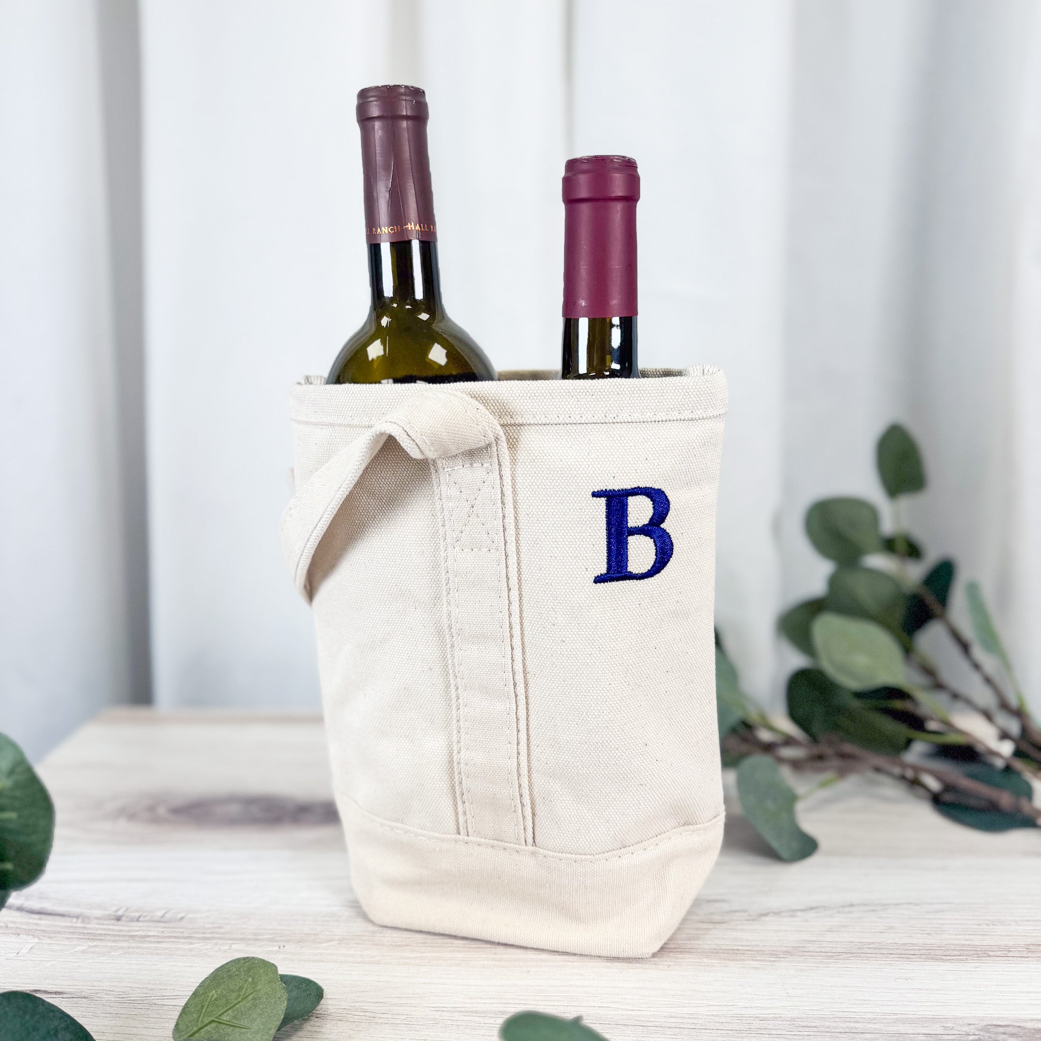Personalized canvas wine tote bag holding two bottles, embroidered with royal blue letter B monogram, styled with eucalyptus on white wood surface