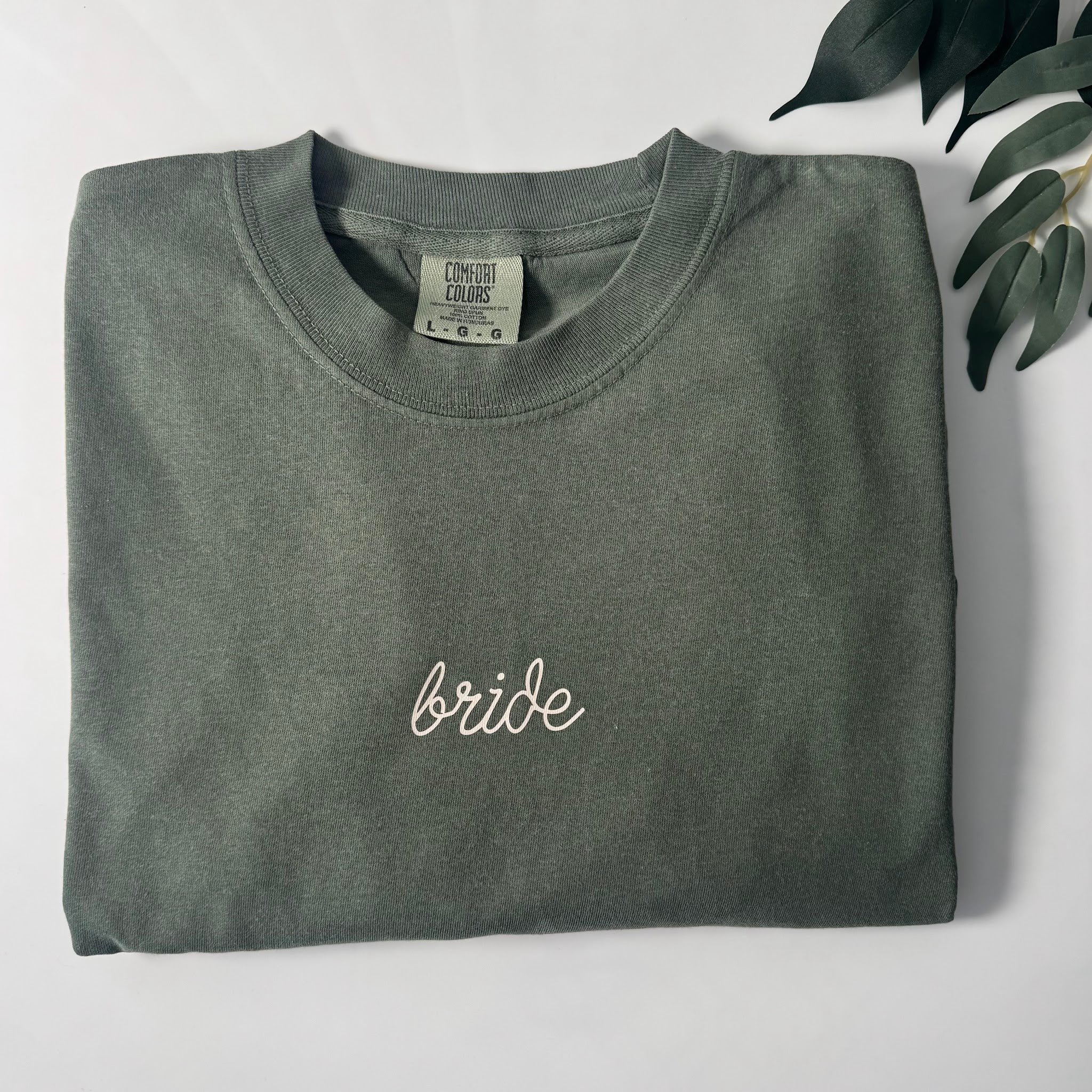 Close-up of white script Bride embroidery on hunter green Comfort Colors 1717 t-shirt