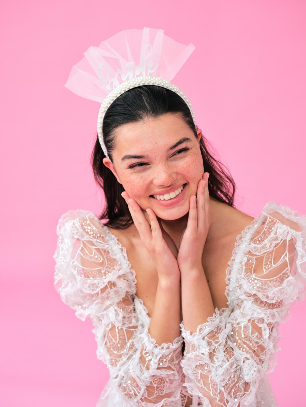 White lace headband with pearl details and sheer tulle bow, worn by smiling woman against pink background.