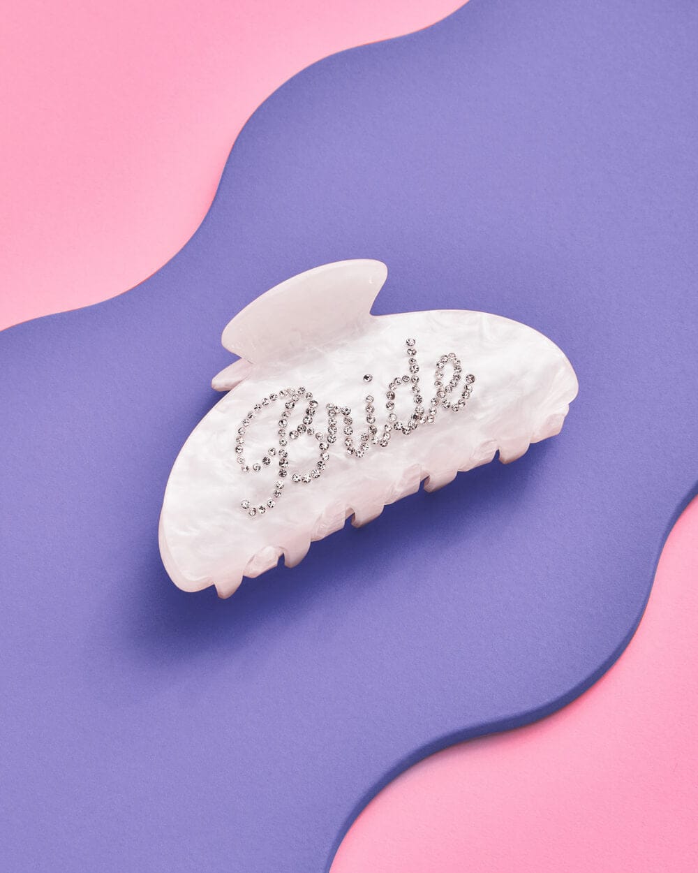 White pearlescent claw clip with "Bride" spelled in rhinestones, displayed on a curved purple and pink background.