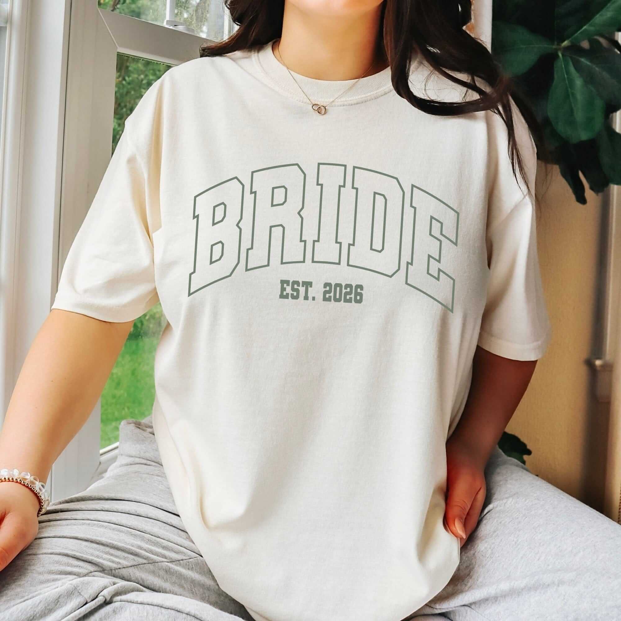 A cream-colored, short-sleeved t-shirt made of soft fabric, featuring a large, outlined collegiate-style graphic, ideal for pre-wedding events.