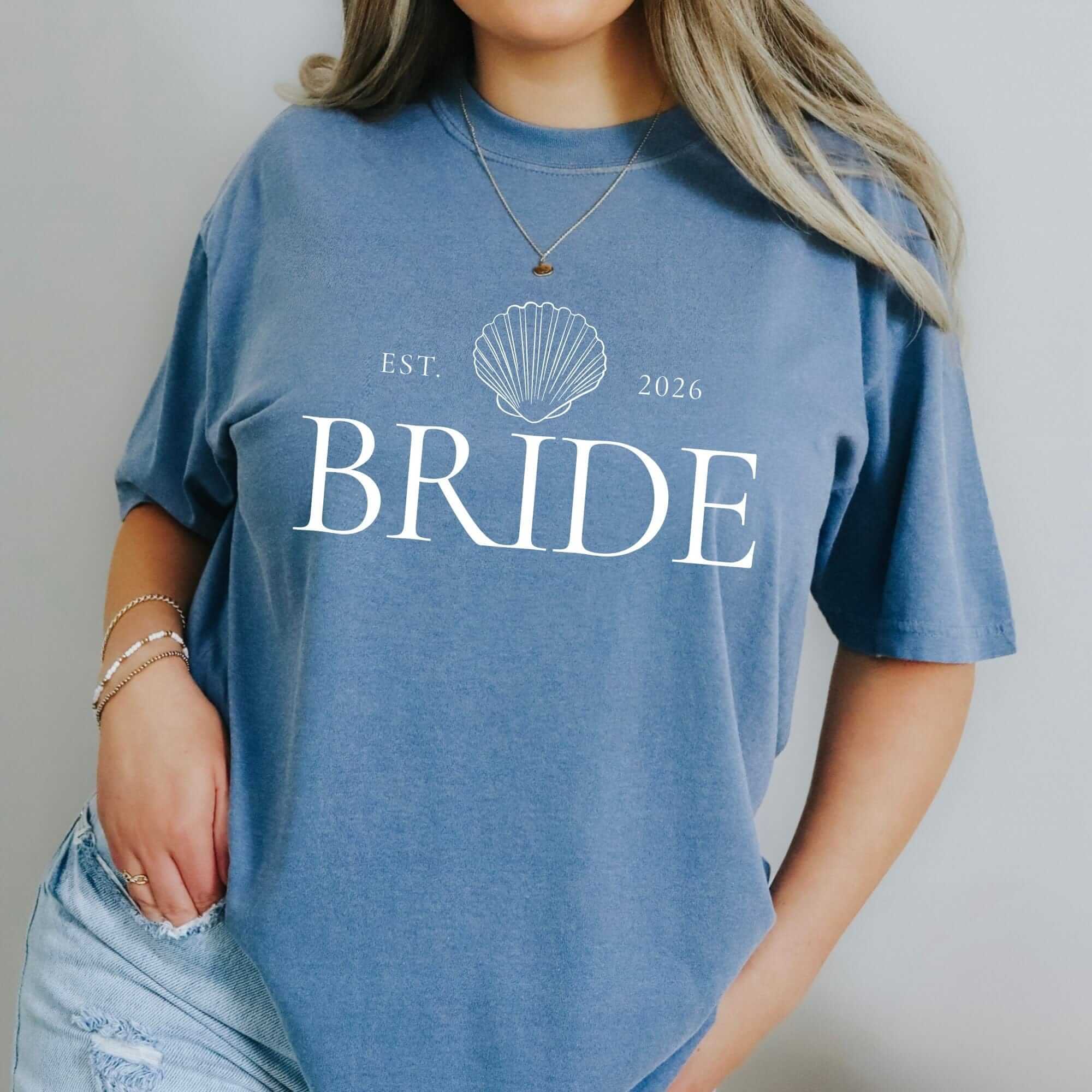 A comfortable periwinkle blue t-shirt made of soft fabric, featuring a white seashell graphic and elegant custom text, perfect for a bridal shower or casual wedding-related wear.