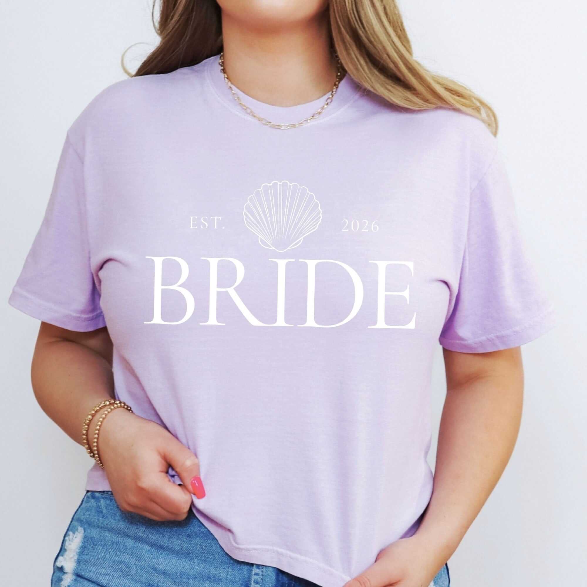 A light purple t-shirt features a white seashell graphic design, perfect for a bridal event or wedding celebration.