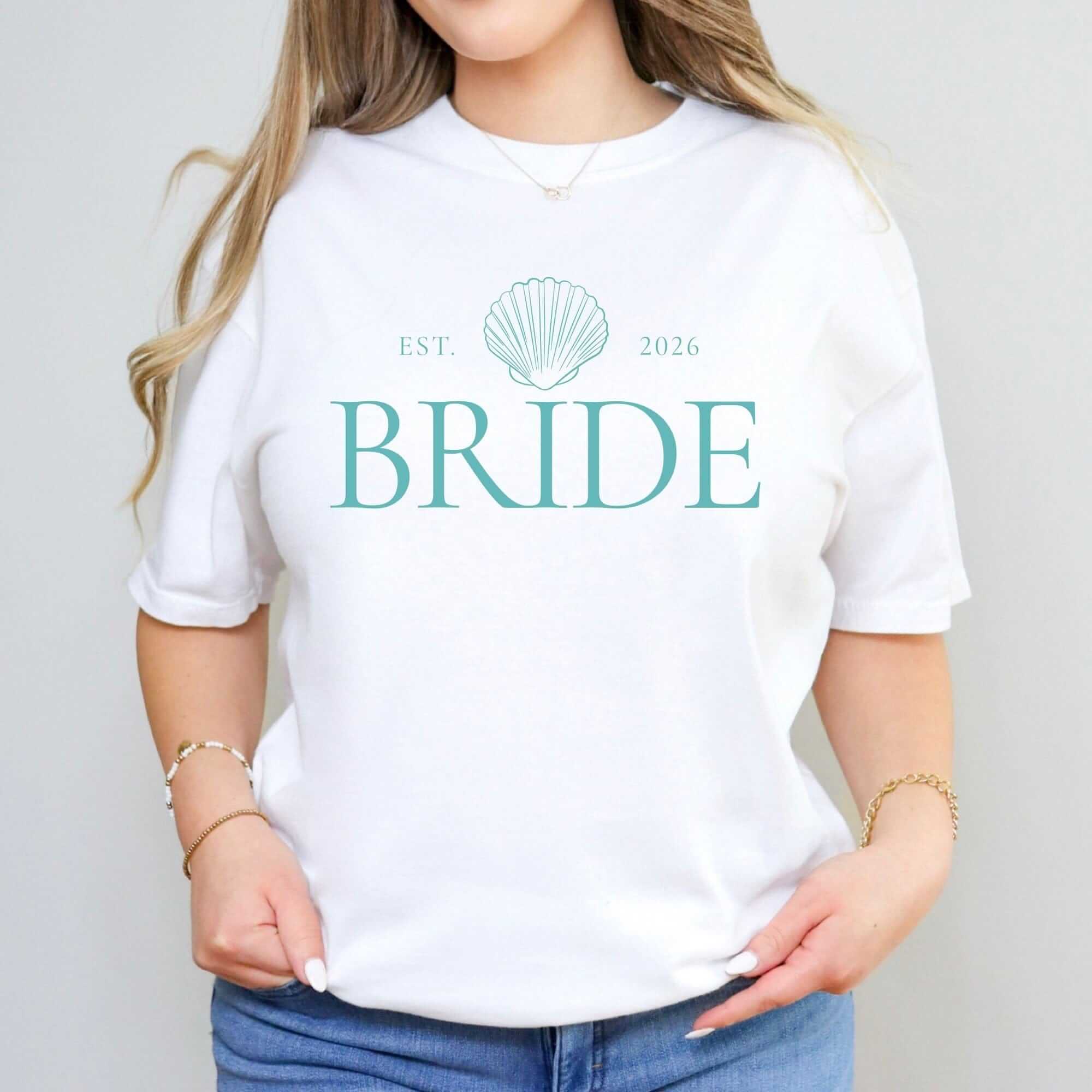 A soft white short-sleeve t-shirt features a prominent teal seashell graphic, ideal for a bridal shower or wedding event.