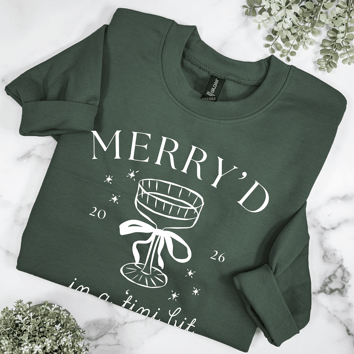 Folded forest green Christmas bride crewneck sweatshirt with Merry'd in a Tini Bit design, white champagne glass graphic, and 2026 date.