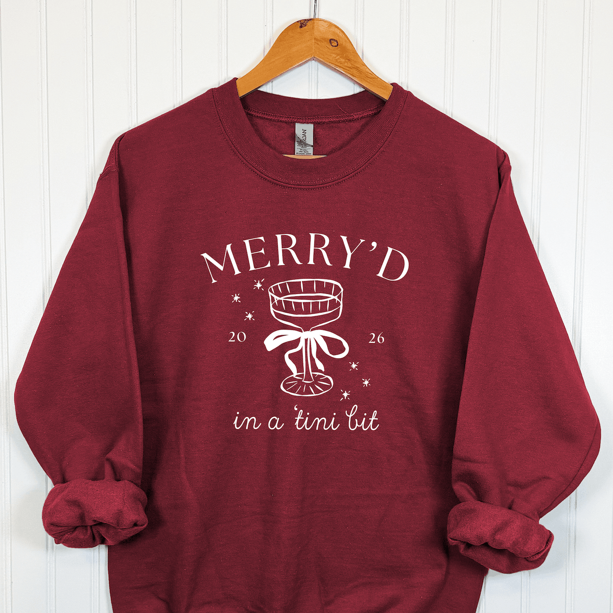 Burgundy Merry'd in a Tini Bit Christmas bride sweatshirt featuring white champagne coupe with bow, festive snowflakes, and holiday bridal lettering.