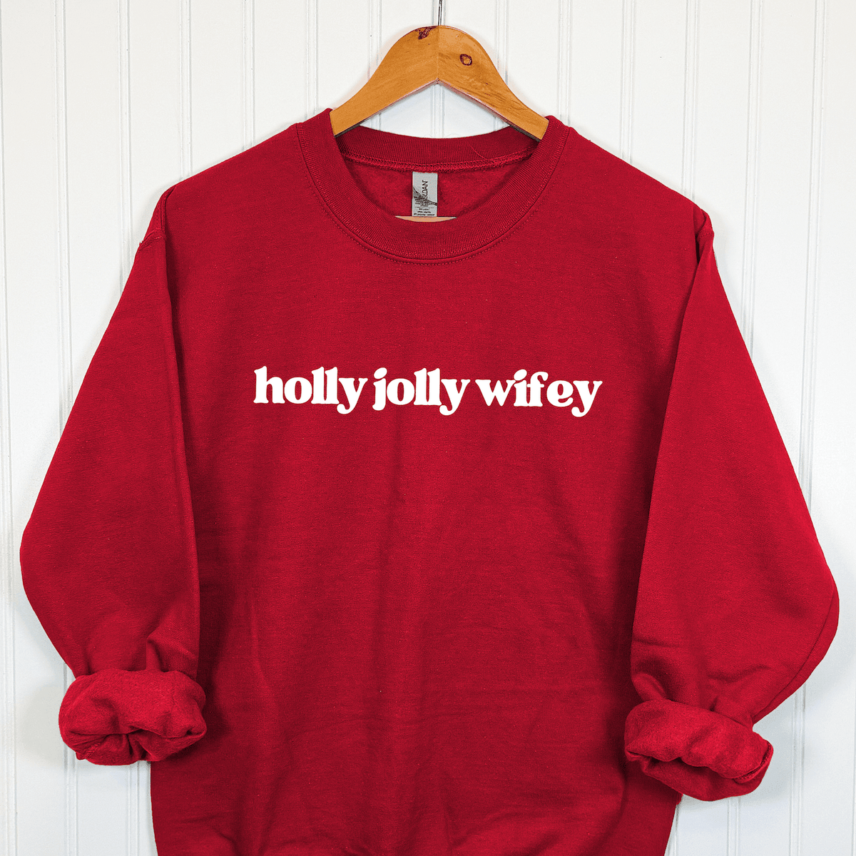 Holly Jolly Wifey Christmas crewneck sweatshirt in festive red with white vintage-inspired lettering, displayed on a wooden hanger against a white wall.