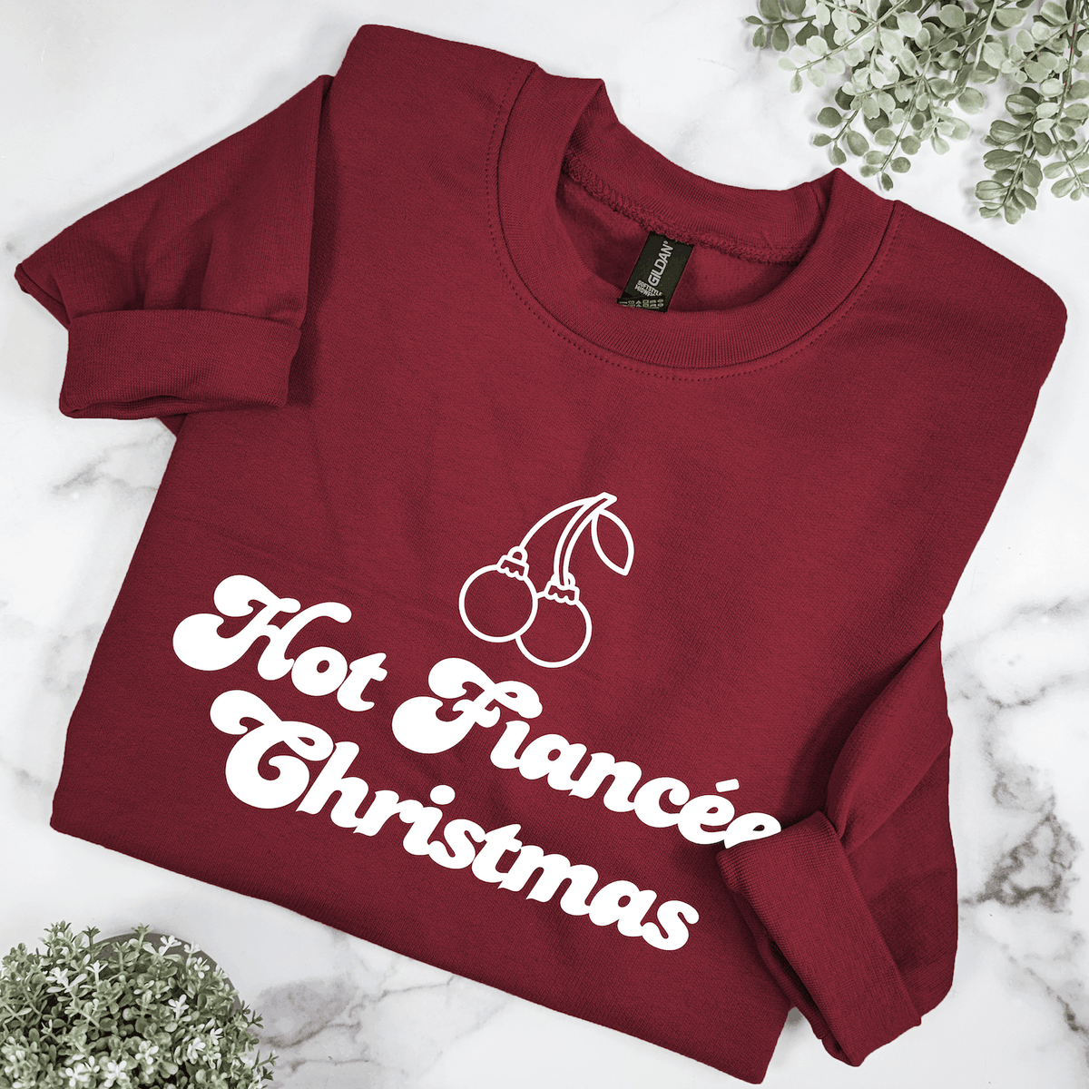 Burgundy Hot Fiancรฉe Christmas sweatshirt featuring white retro lettering and cherry ornament illustration, folded on a marble background with greenery.