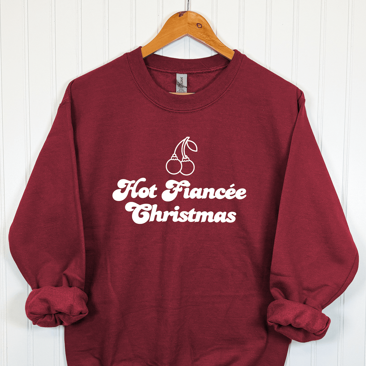 Hot Fiancée Christmas bride crewneck sweatshirt in burgundy with white vintage-inspired lettering and cherry Christmas ornaments, displayed on a wooden hanger.