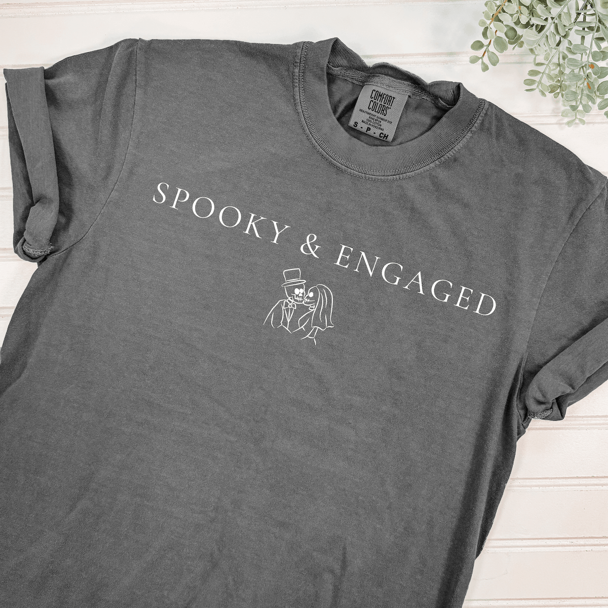 Gray Spooky & Engaged Halloween bride t-shirt featuring white lettering and skeleton bride and groom illustration, styled flat with greenery.