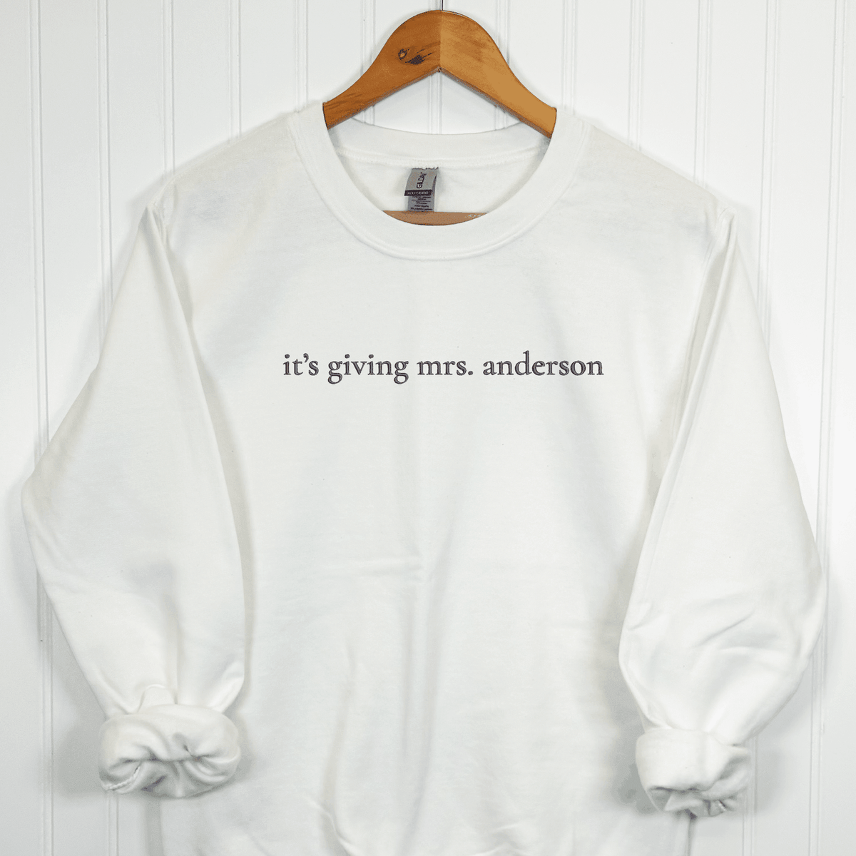White personalized It's Giving Mrs. embroidered bride sweatshirt featuring custom last name in dark gray thread, displayed on a wooden hanger.