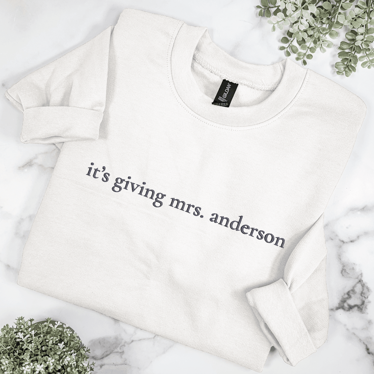 Folded white It's Giving Mrs. bridal crewneck sweatshirt with personalized last name embroidery in dark gray, styled on a marble background with greenery.