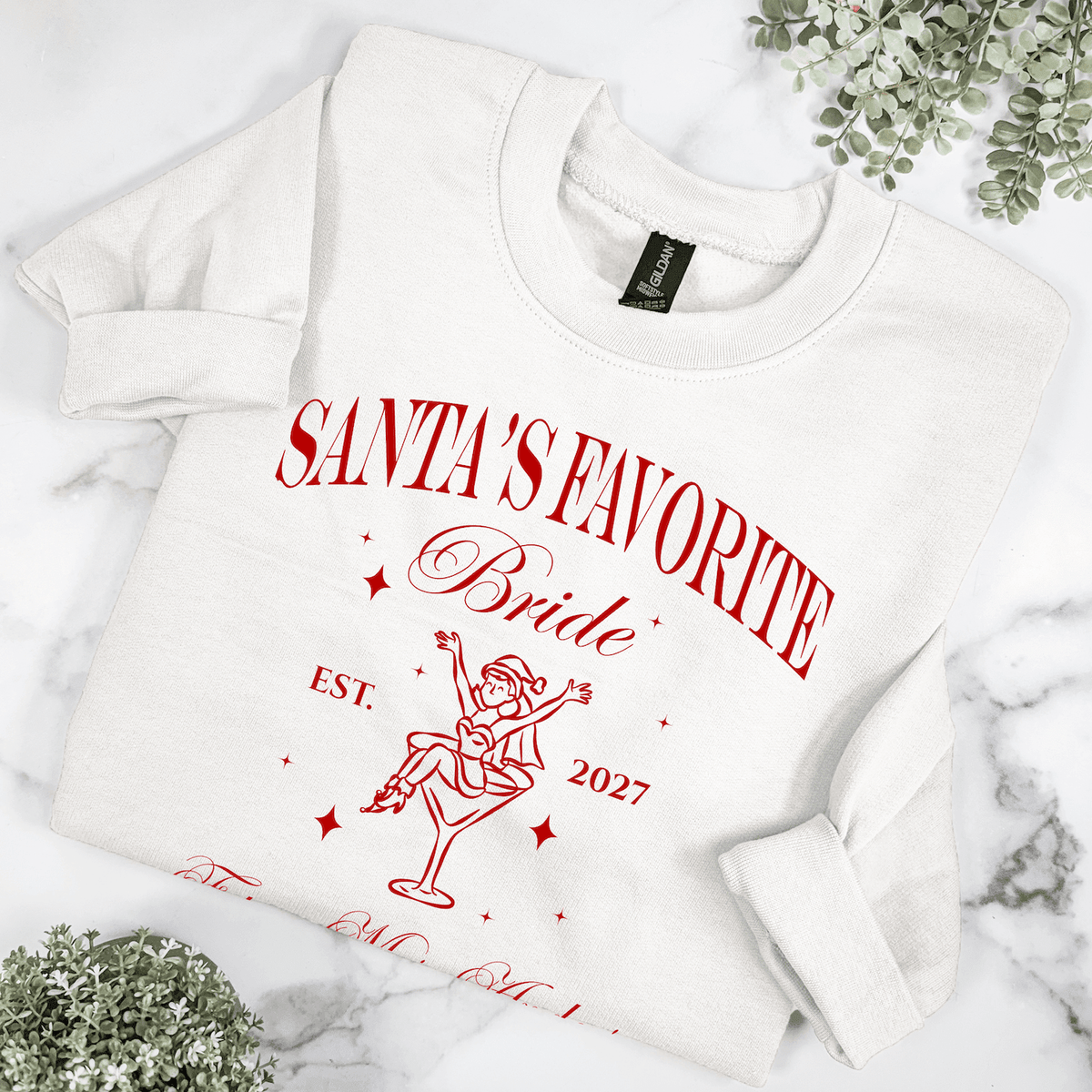 Close-up of white Santa's Favorite Bride crewneck sweatshirt with red Christmas illustration, personalized bride name and wedding year.