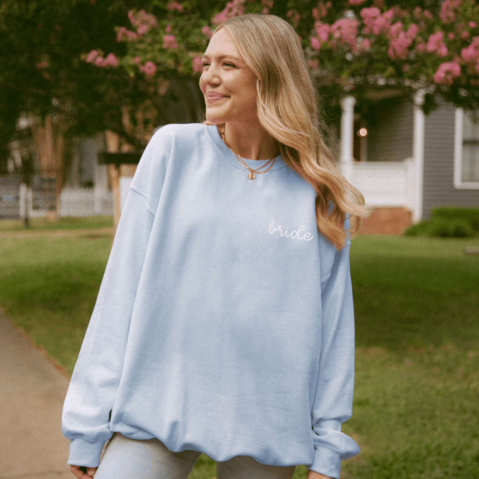 Light blue crewneck sweatshirt with embroidered "Bride" text on chest, worn outdoors with pink flowering bushes in background.