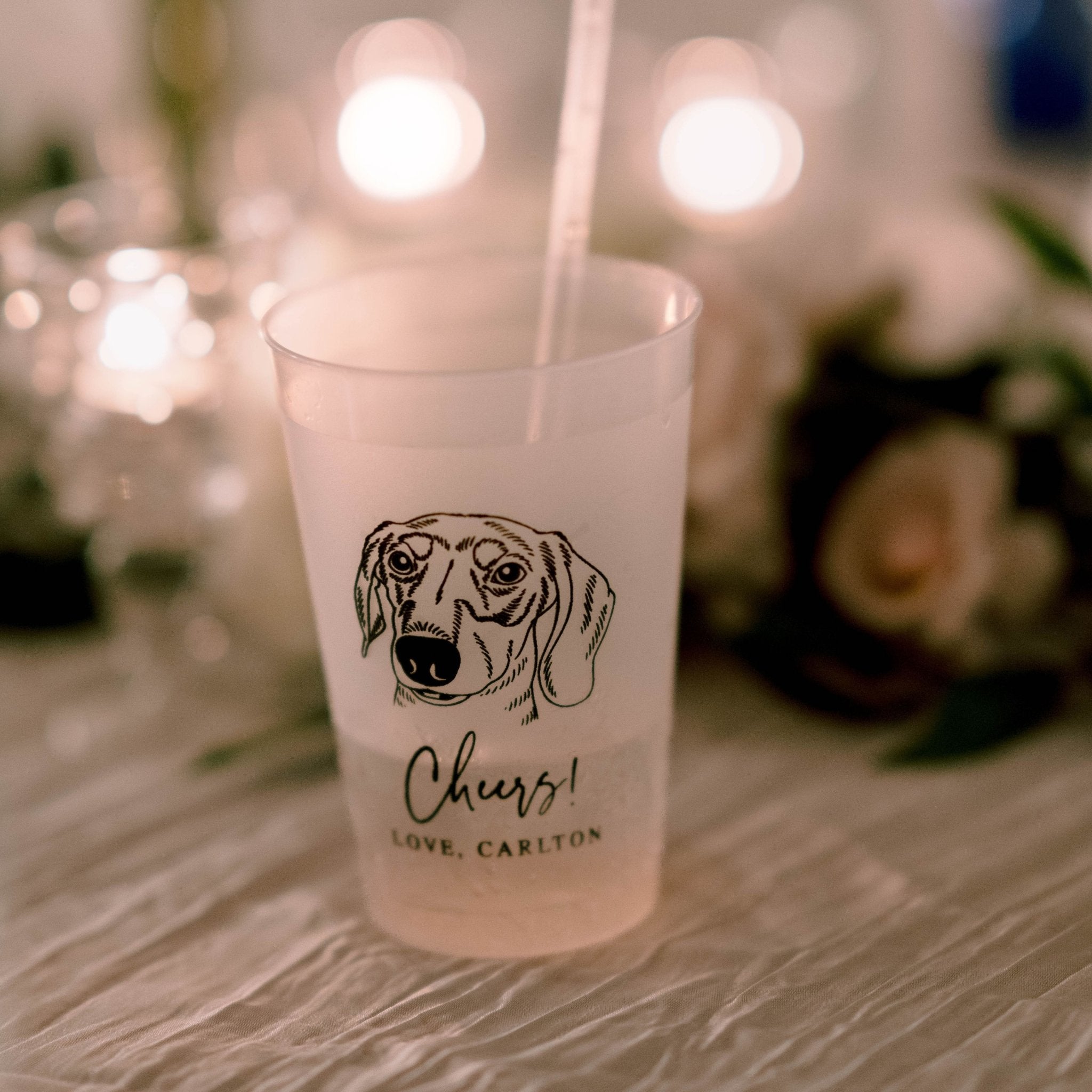 Frosted plastic cup with dog illustration and "Cheers! Love, Carlton" personalization text, displayed with string lights and floral background.