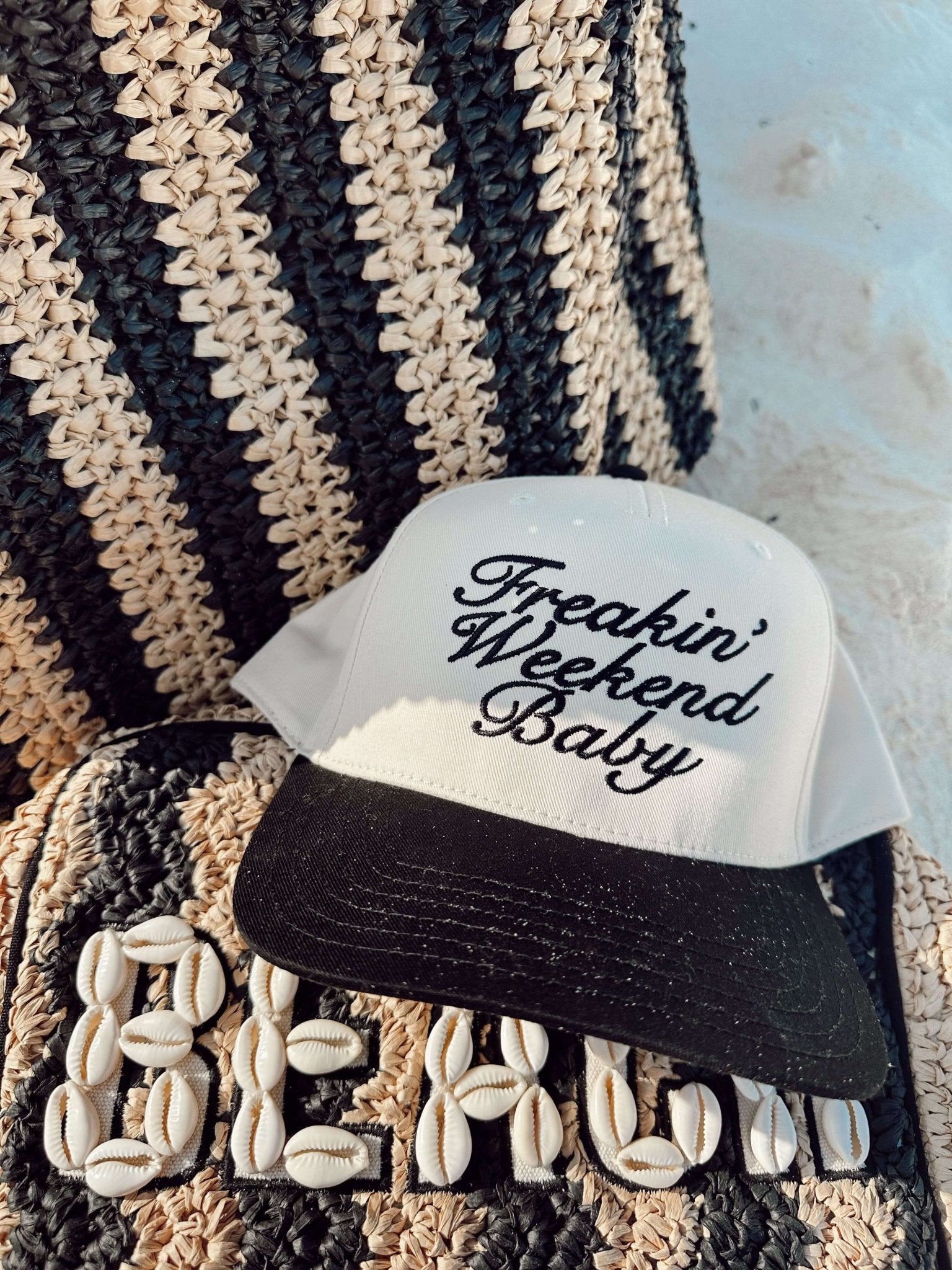 Black and white vintage trucker hat with "Freakin' Weekend Baby" script embroidery on the front panel, displayed on striped fabric background.