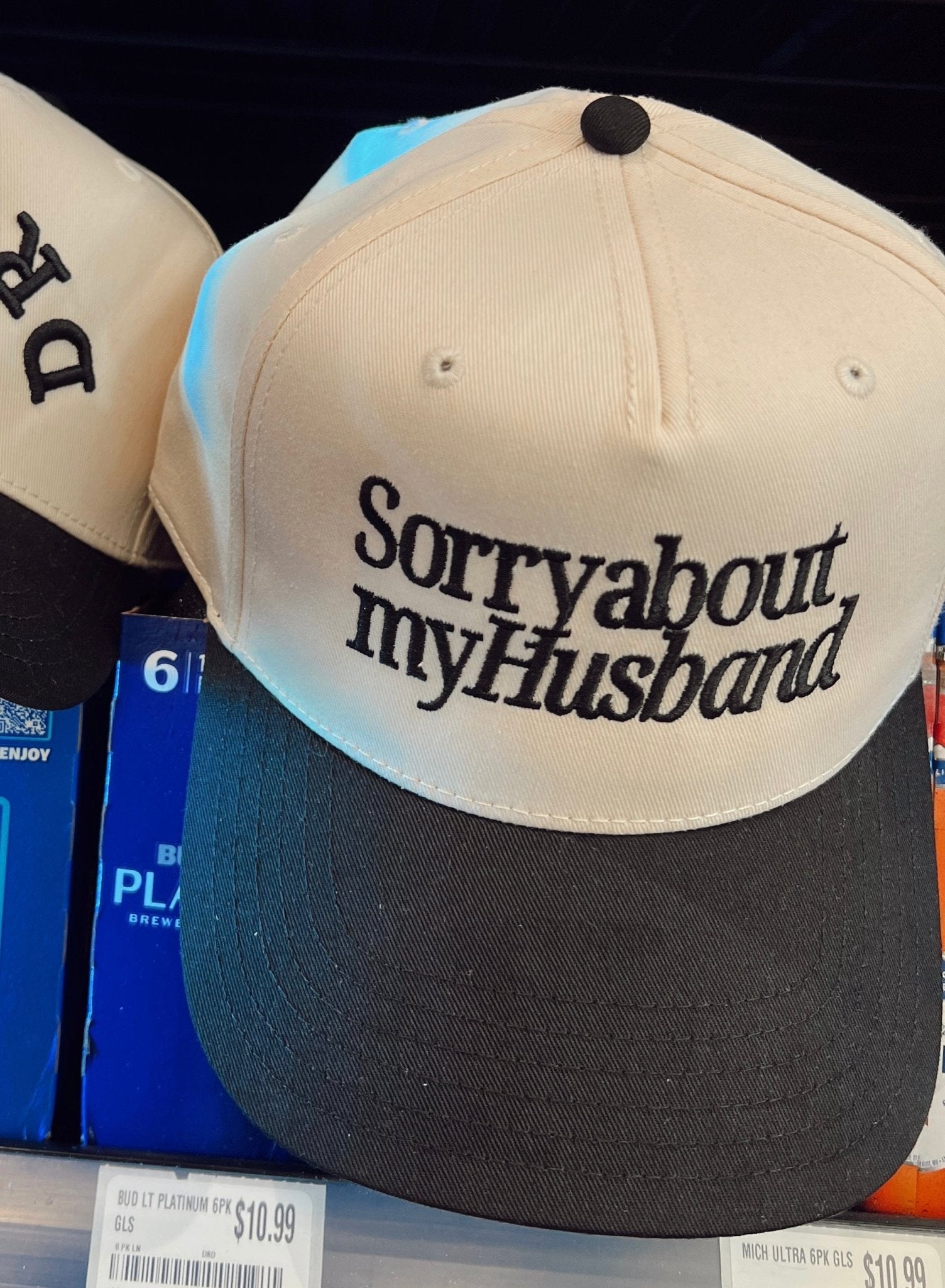 Cream and charcoal trucker hat with "Sorry about my Husband" embroidered across the front in black text