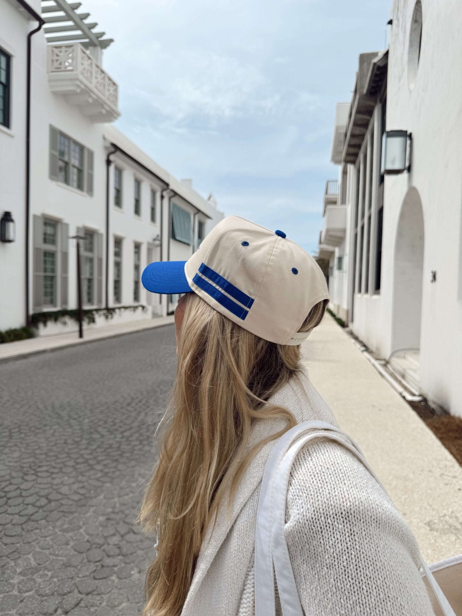 Cream and royal blue vintage trucker hat with striped detail, worn by woman on cobblestone street lined with white buildings.