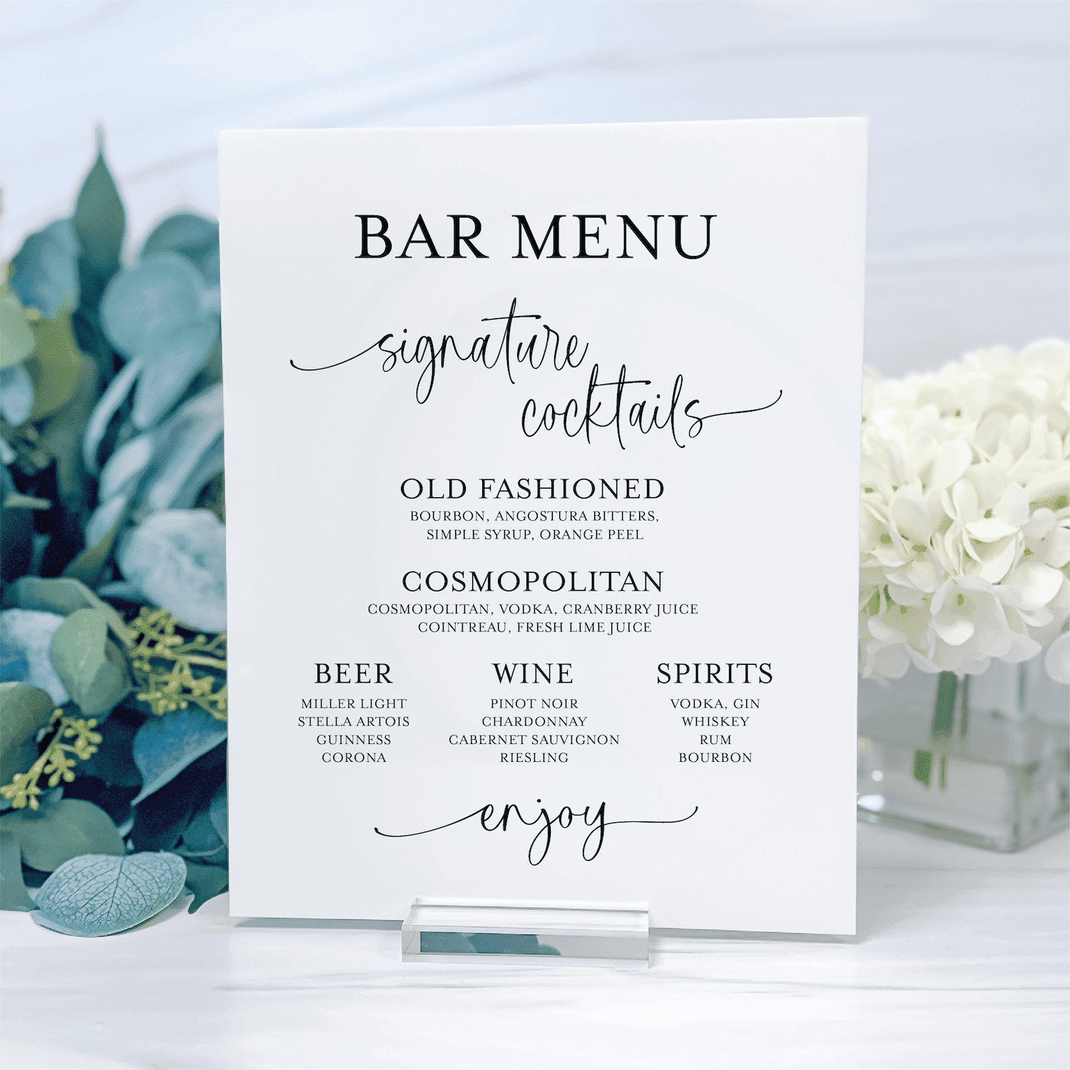 White acrylic bar menu sign displaying signature cocktails, beer, wine, and spirits options with elegant black script and serif typography, displayed on a stand with greenery and white flowers.