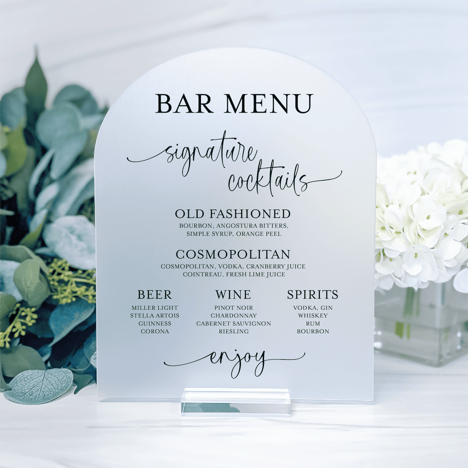 White acrylic bar menu sign displaying signature cocktails, beer, wine, and spirits with elegant script and serif typography, surrounded by greenery and white flowers.
