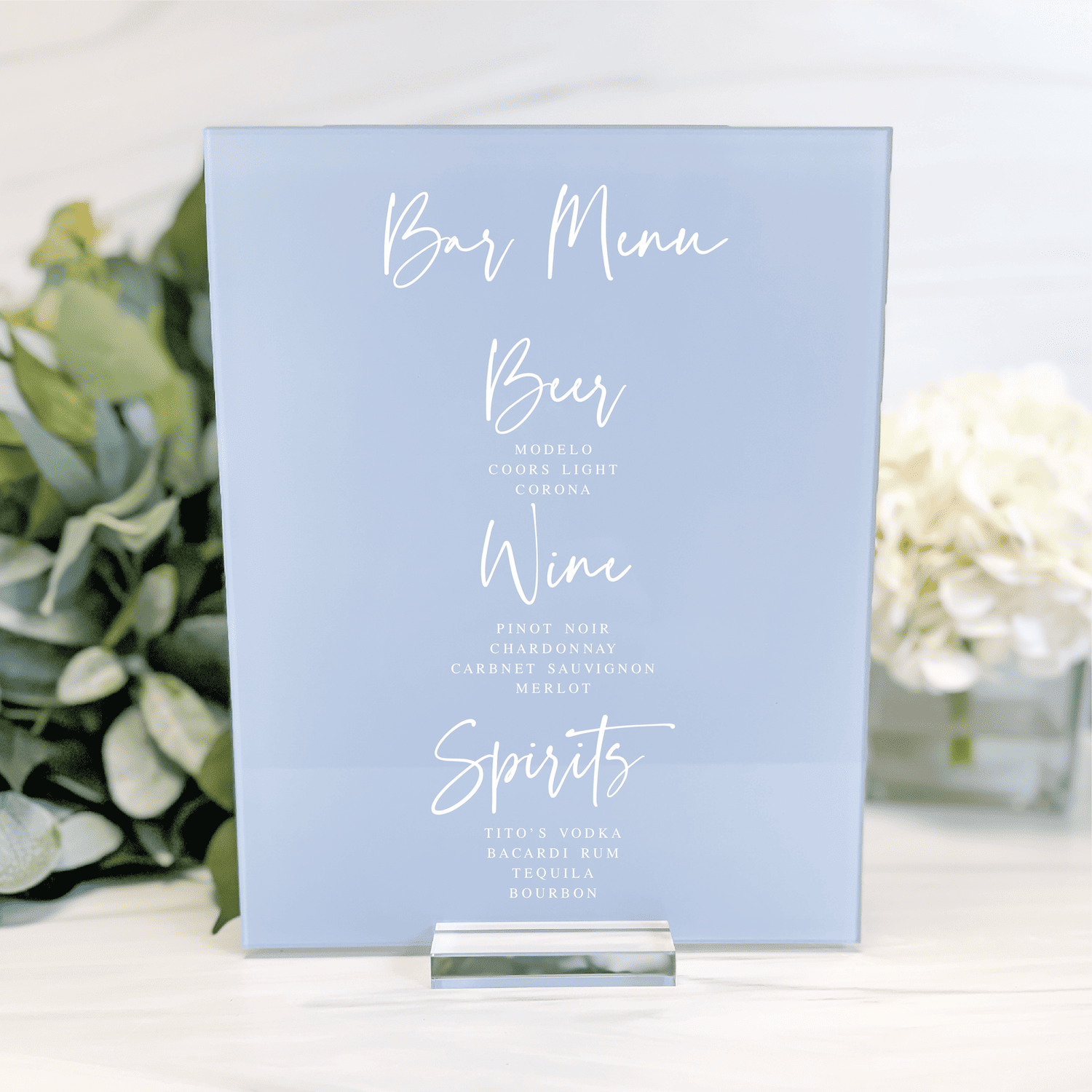 Light blue bar menu sign with white script text listing Beer, Wine, and Spirits options, displayed on a stand with greenery beside it.