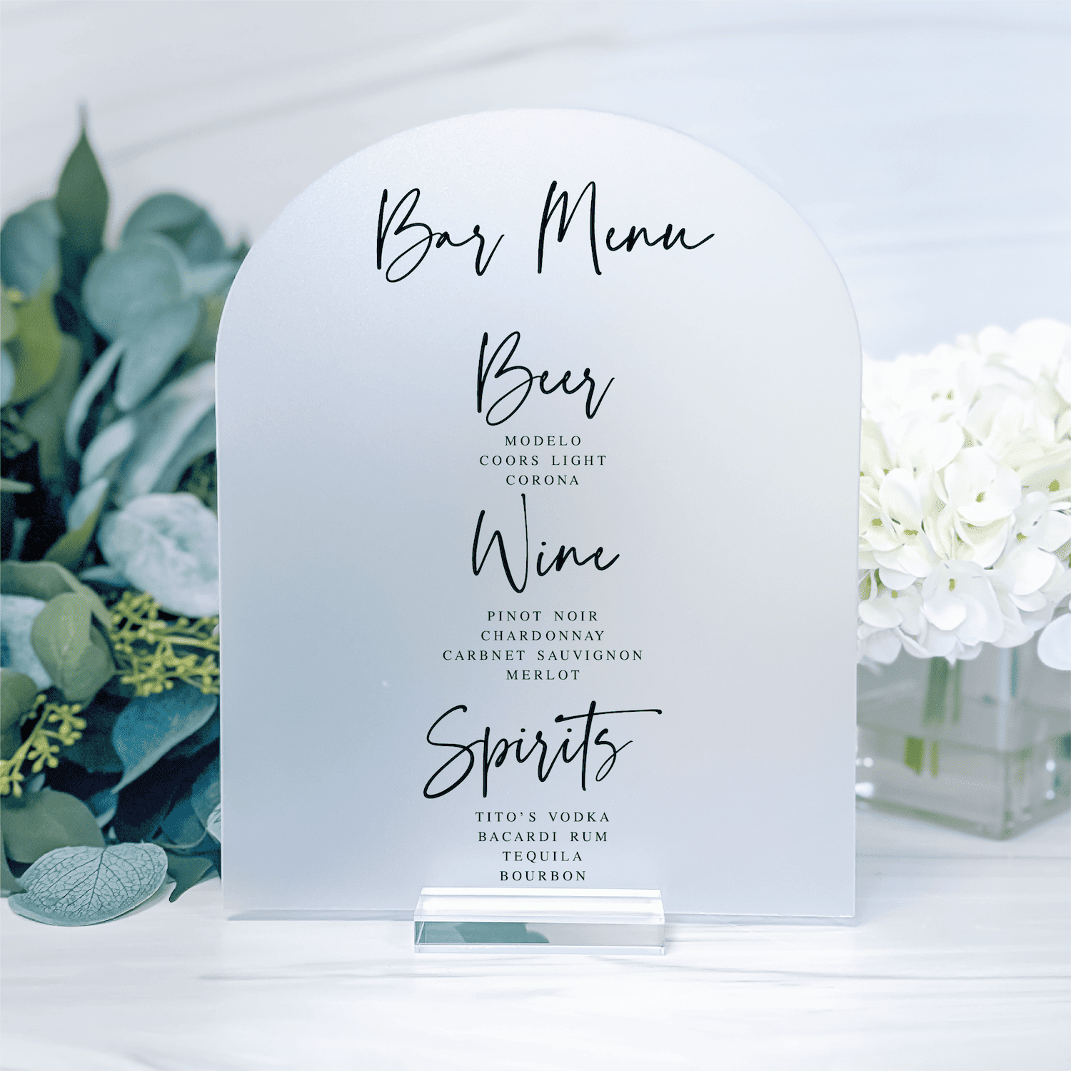 White arch-shaped bar menu sign with handwritten-style text listing Beer, Wine, and Spirits options, flanked by greenery and white flowers.