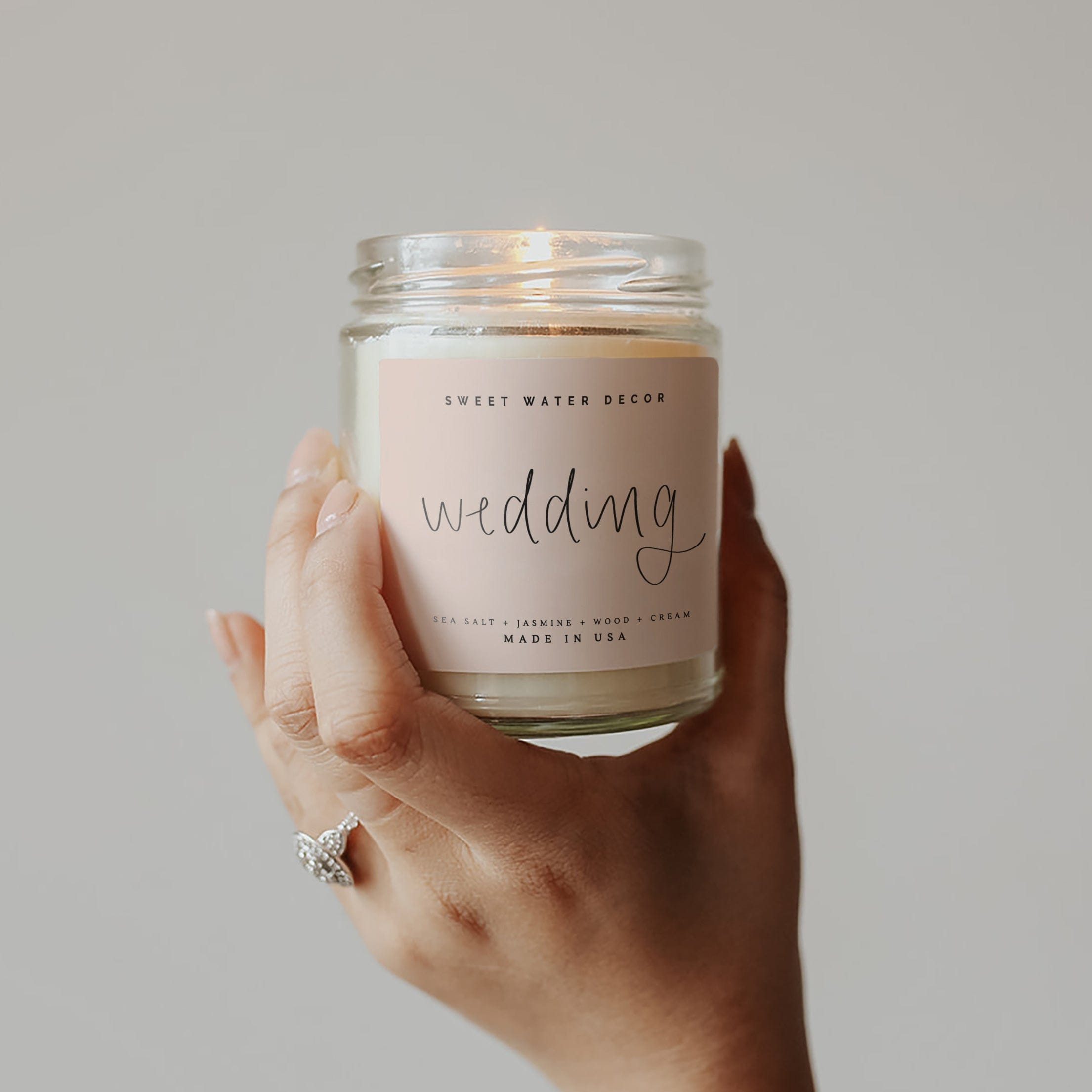 Hand holding clear glass jar candle with "wedding" text on cream label, Sweet Water Decor brand