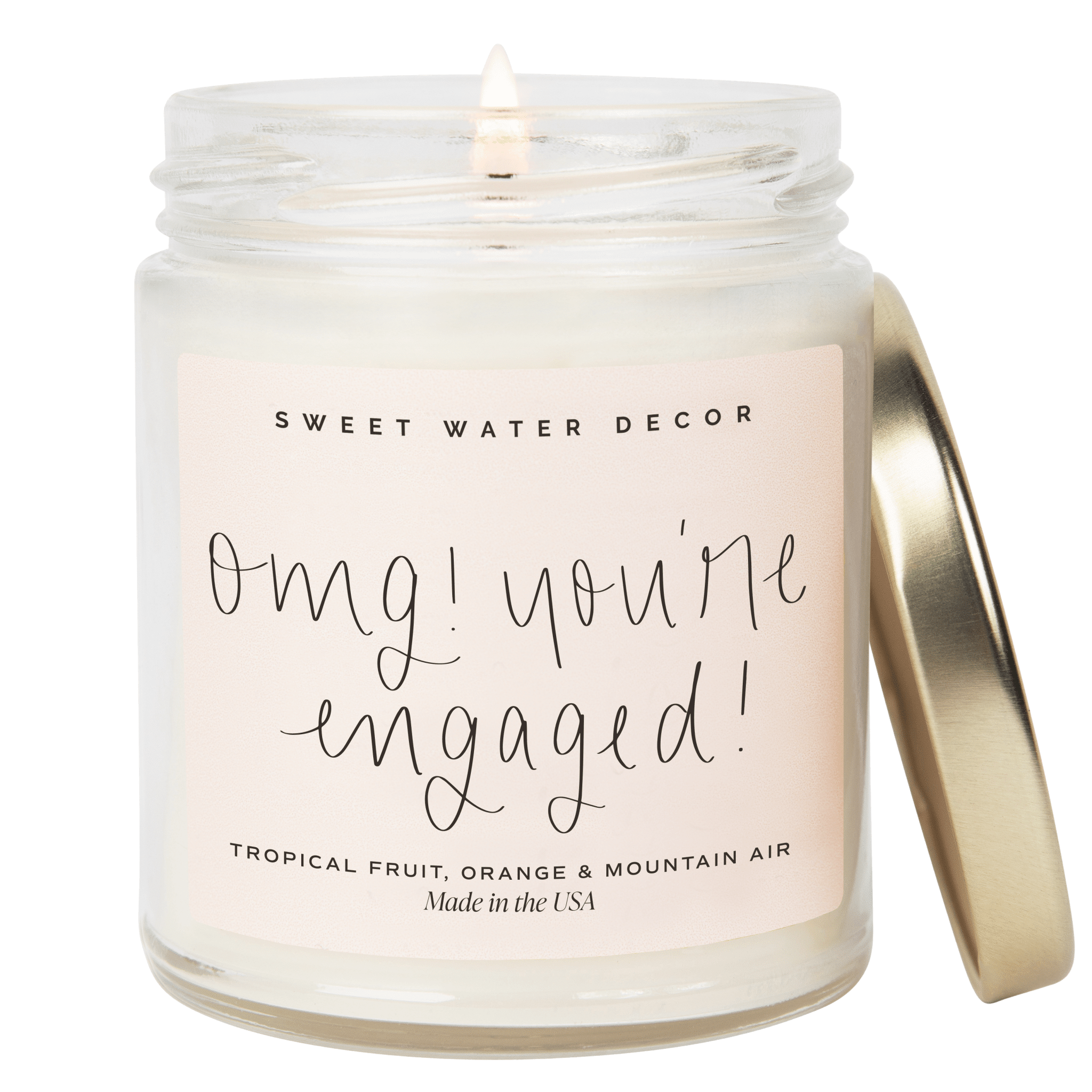 White soy candle in clear glass jar with gold lid, labeled "OMG! you're engaged!" in script lettering and "Sweet Water Decor" branding.