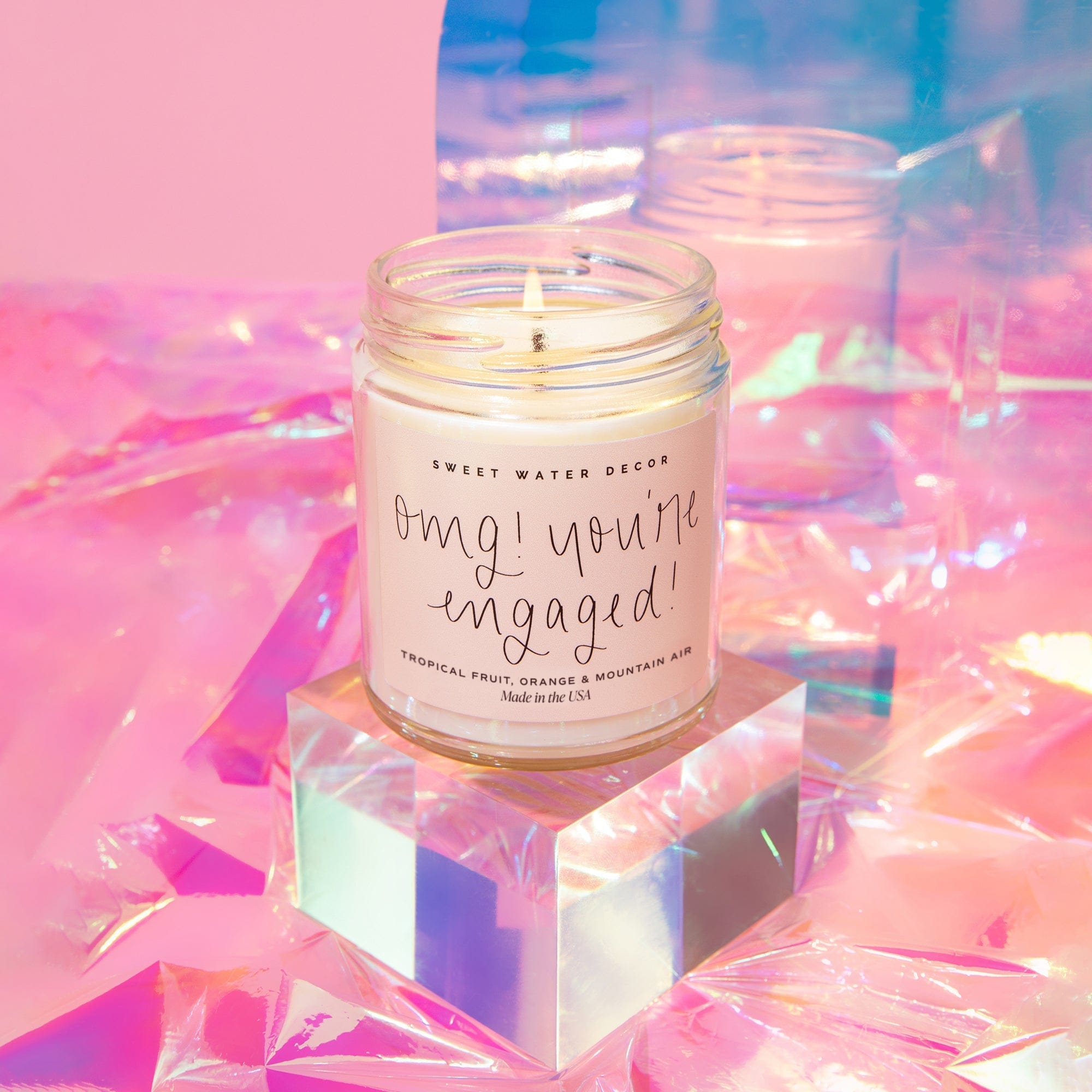 Cream soy candle in clear glass jar with "omg! you're engaged!" text, displayed on iridescent cube with pink and blue lighting.