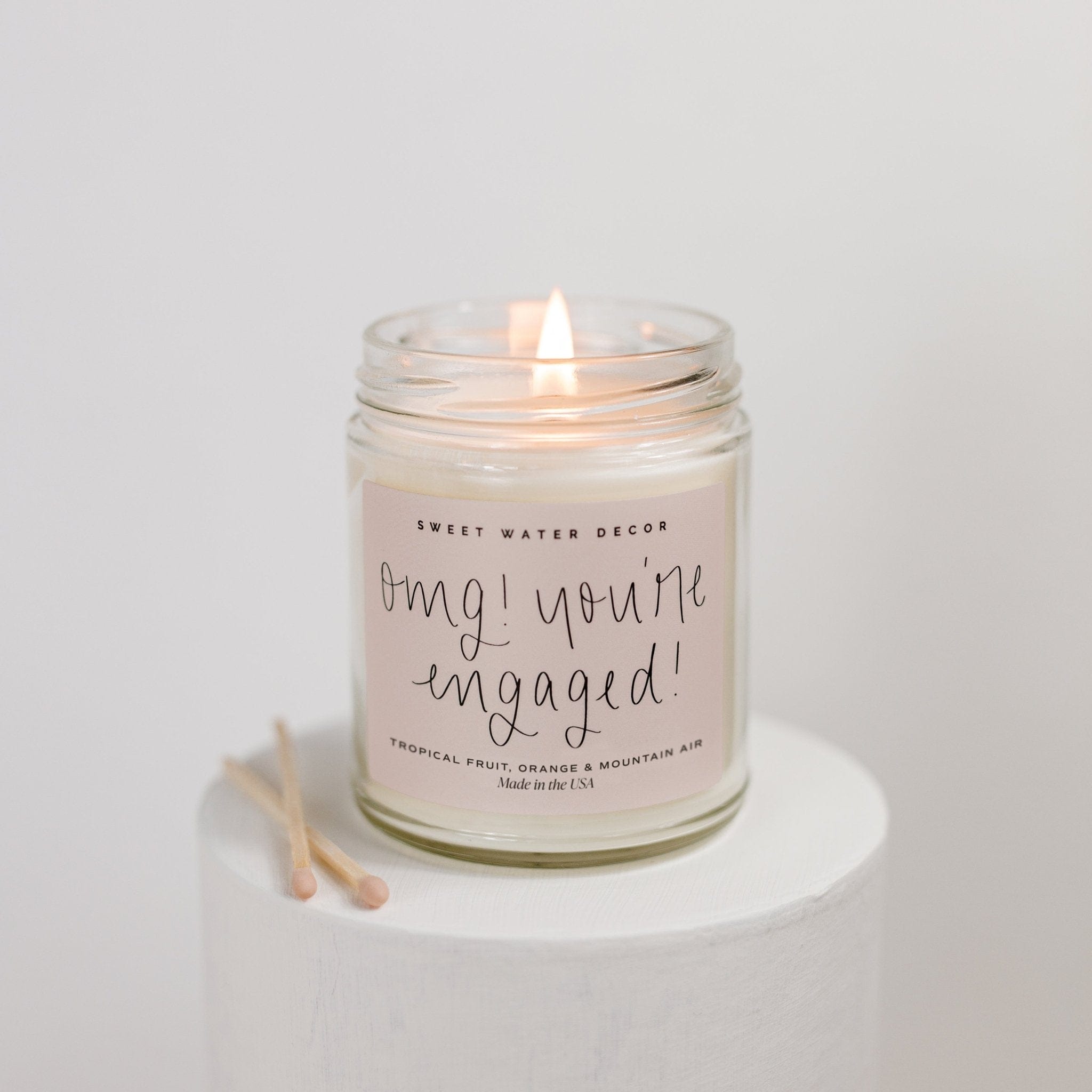 Lit soy candle in clear glass jar with "OMG you're engaged" script text, sitting on white surface with matches.