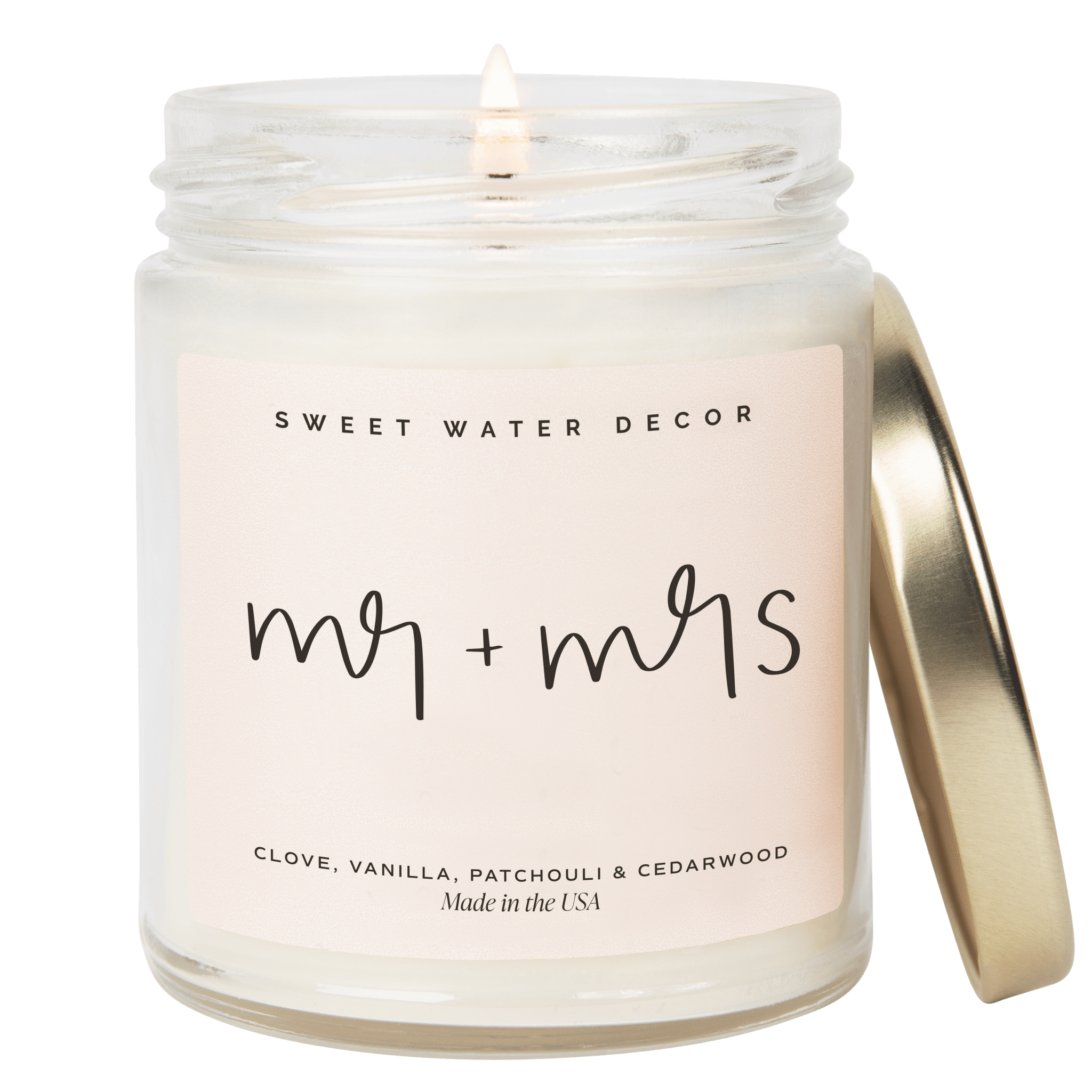 Clear glass jar candle with "my + my's" handwritten text, gold lid slightly ajar, lit wick visible inside, Sweet Water Decor branding at top.