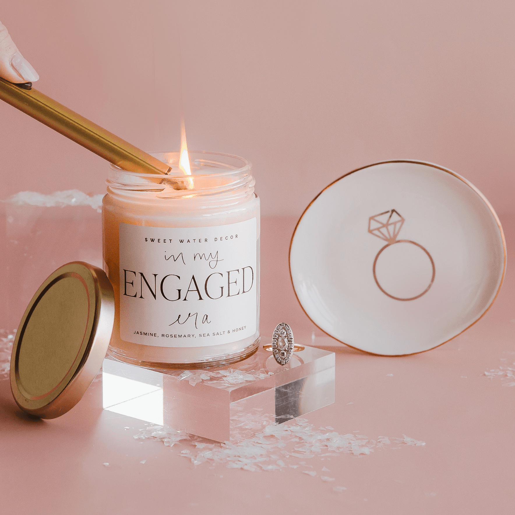 Lit soy candle in clear glass jar with "In My Engaged Era" text, gold lid, matching ring-design dish, and wooden wick on pink background.
