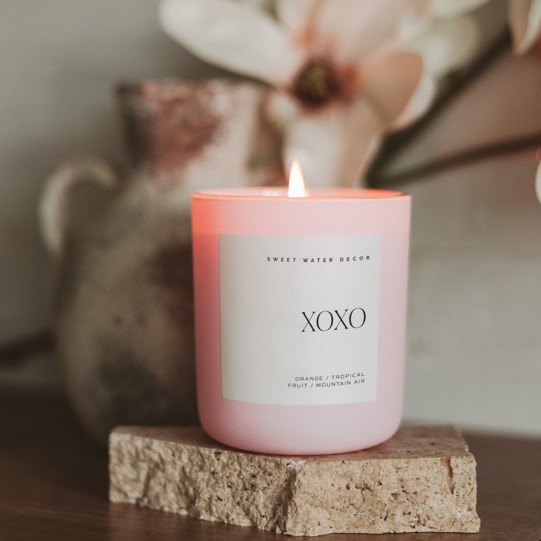 Pink matte jar candle with "XOXO" text and lit wick, displayed on textured stone base with dried flowers