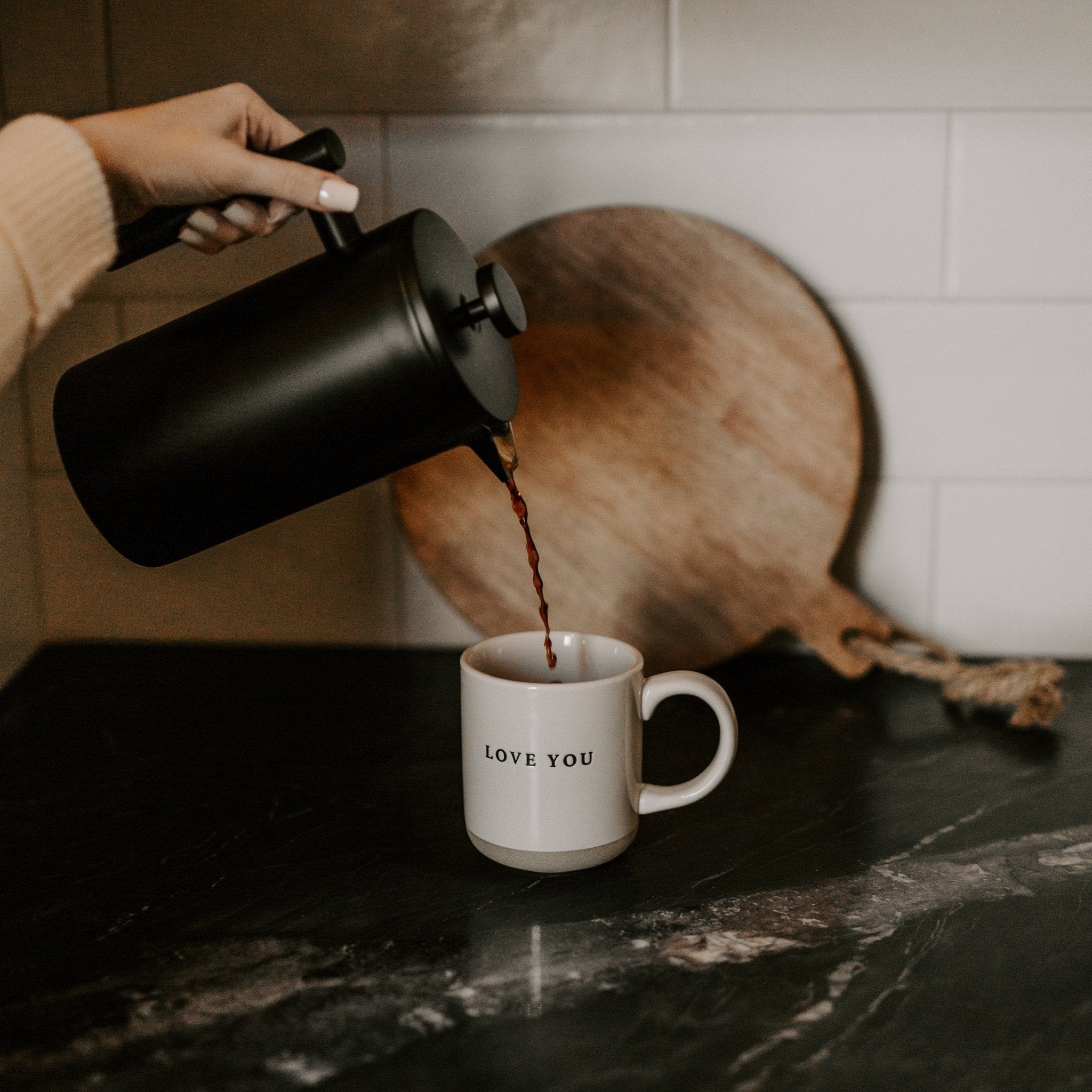 Hand pouring coffee into white ceramic mug with "LOVE YOU" text, black kettle and wooden cutting board visible.