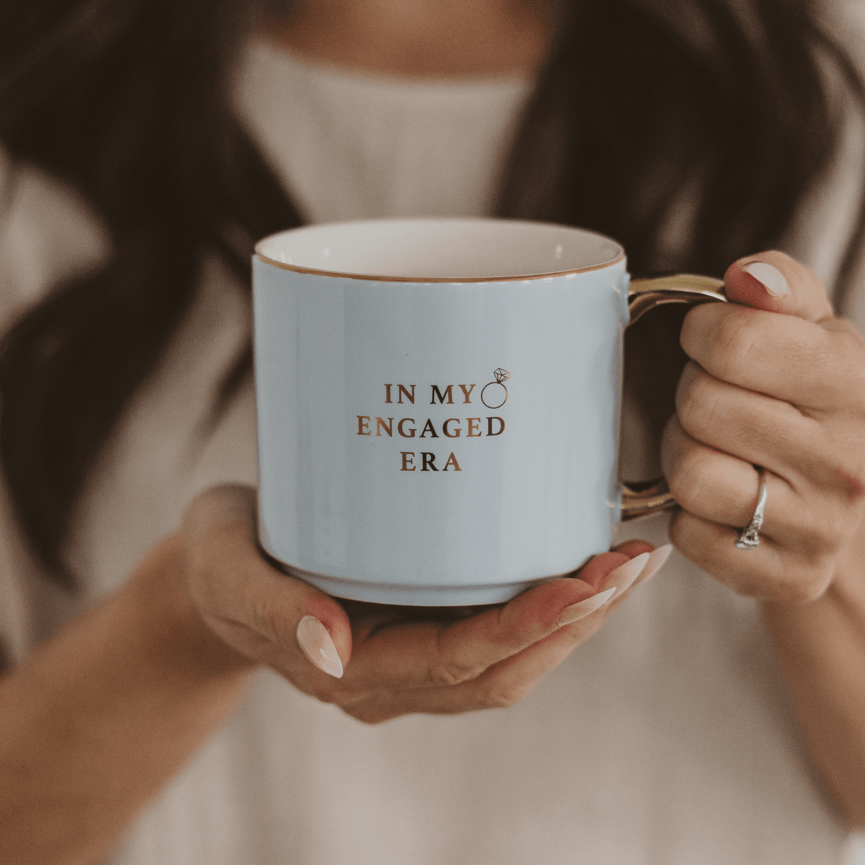 Light blue ceramic mug with gold handle and "IN MY ENGAGED ERA" text, held by woman wearing engagement ring.