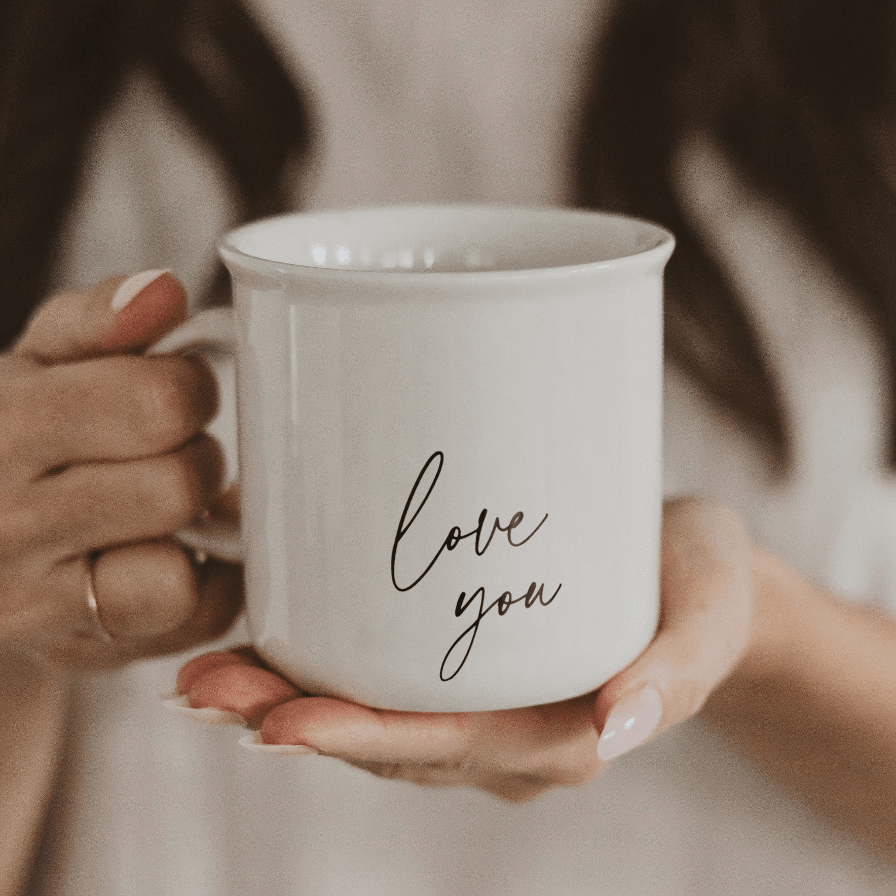 White ceramic mug with "love you" in script text, held in hands against a neutral beige sweater background.