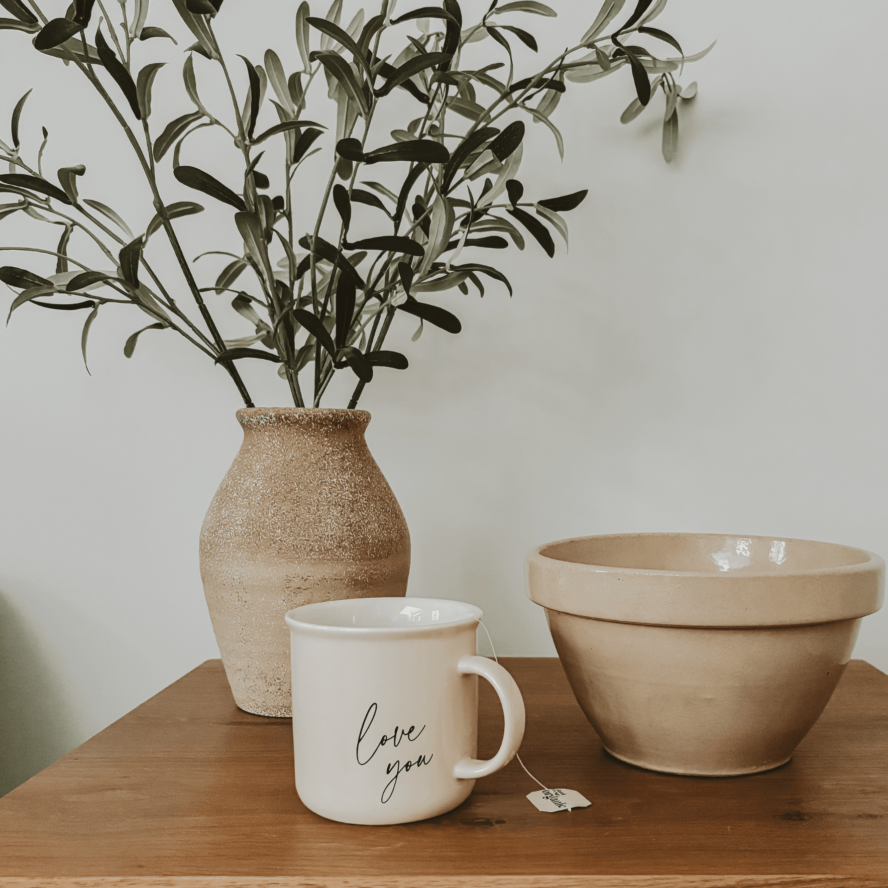 White ceramic coffee mug with "Love you" script text, displayed on wood table next to beige bowls and cork vase with greenery.