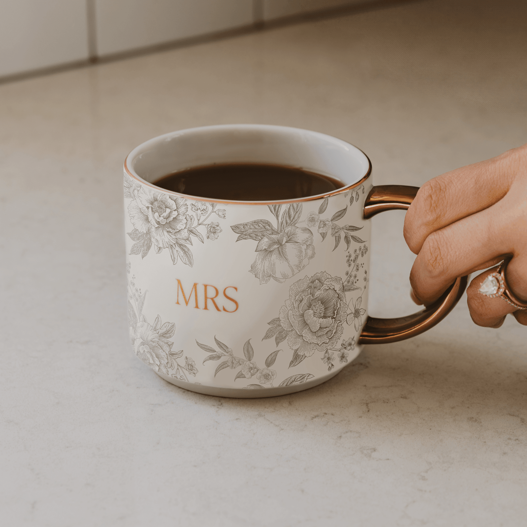 Cream ceramic mug with gold handle, delicate floral pattern, and gold "MRS" text, filled with coffee