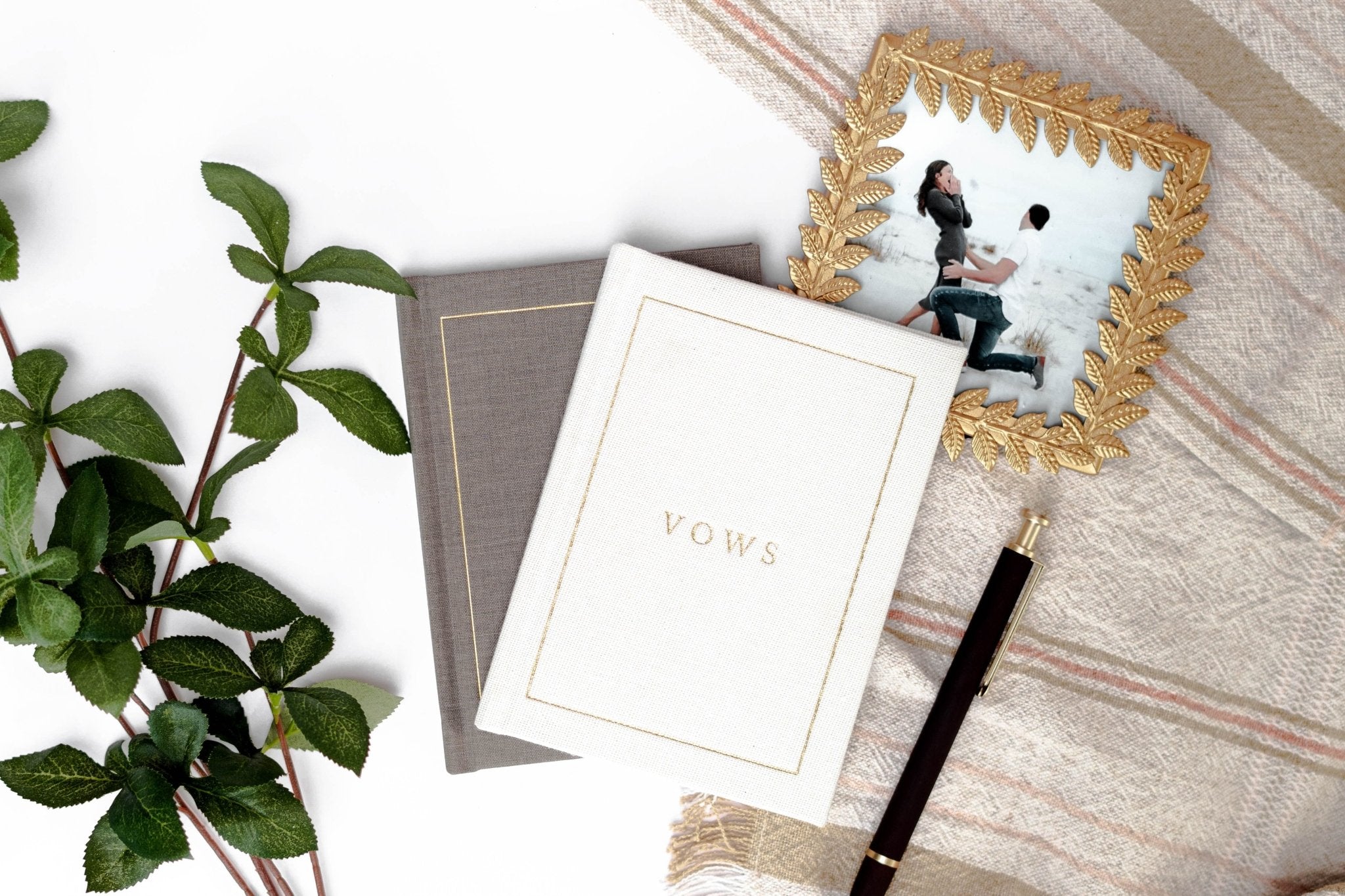 White vow book with gold embossed text next to gray cover, styled with green plant, pen, and framed photo on wooden surface.