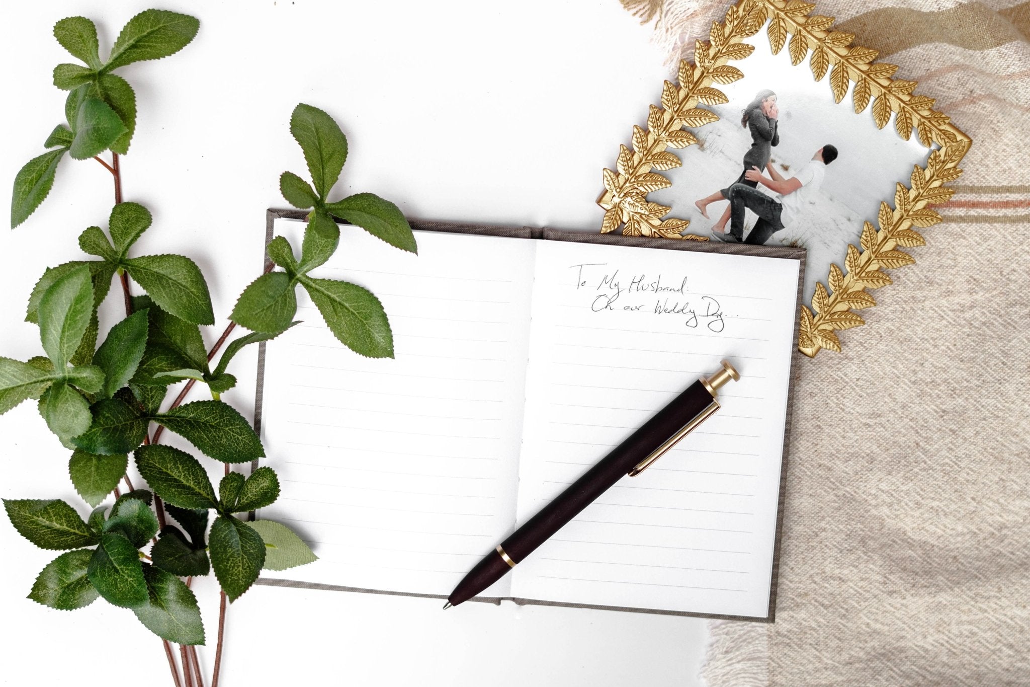 Black pen resting on open white vow book next to fresh green plant and framed photo on neutral fabric background.
